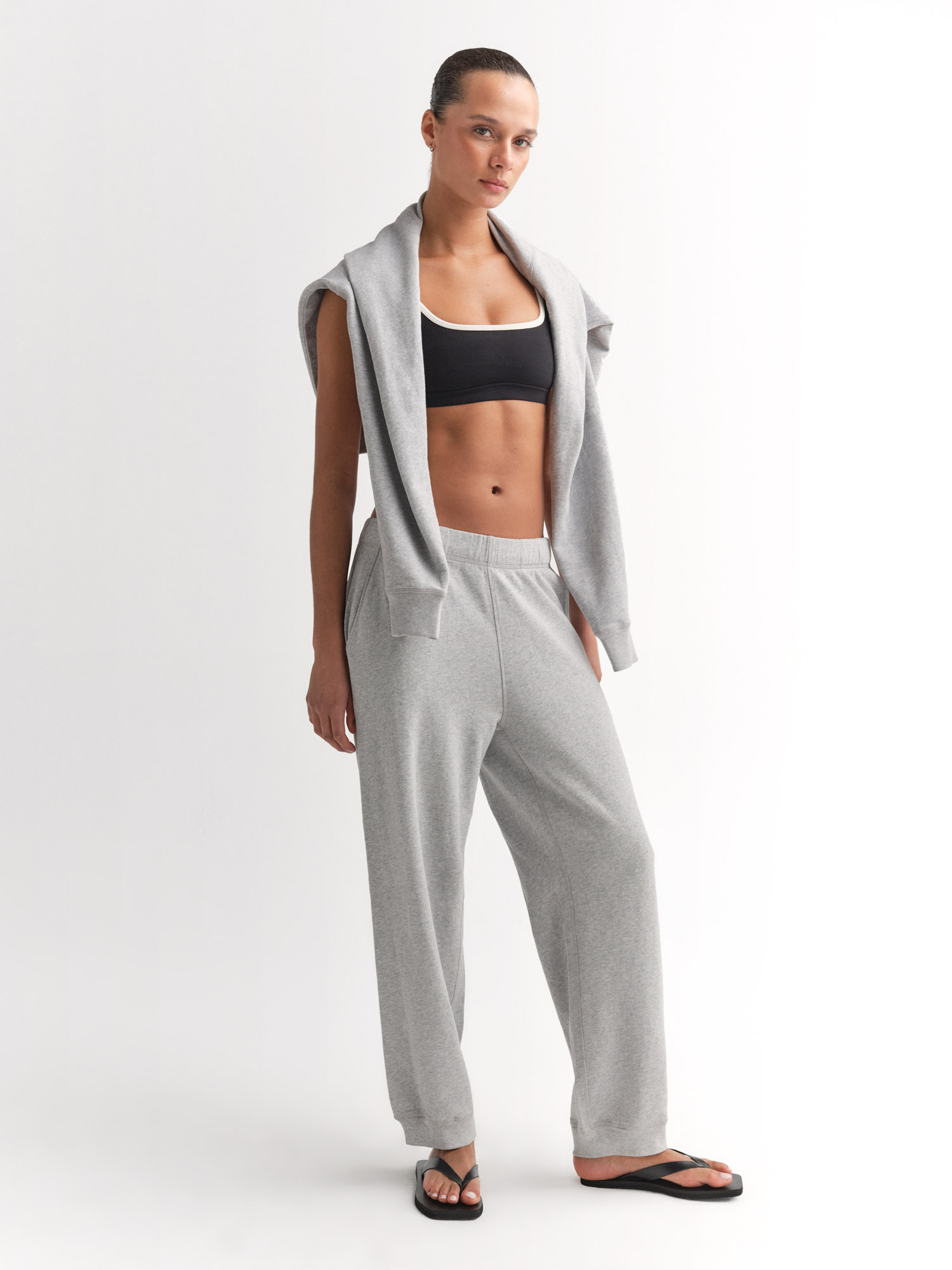 A person wearing a black sports bra, gray sweatpants, a gray sweatshirt draped over their shoulders, and black flip-flops stands against a plain white background.