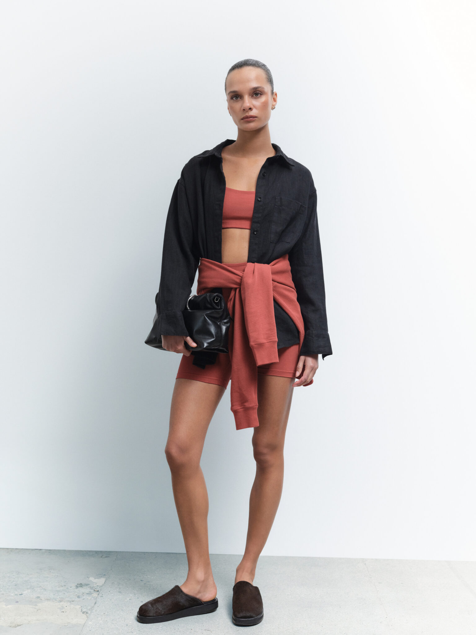 A woman stands against a white background wearing a black button-up shirt, rust-colored crop top and shorts, matching sweatshirt tied at her waist, dark slip-on shoes, and holding a black clutch.