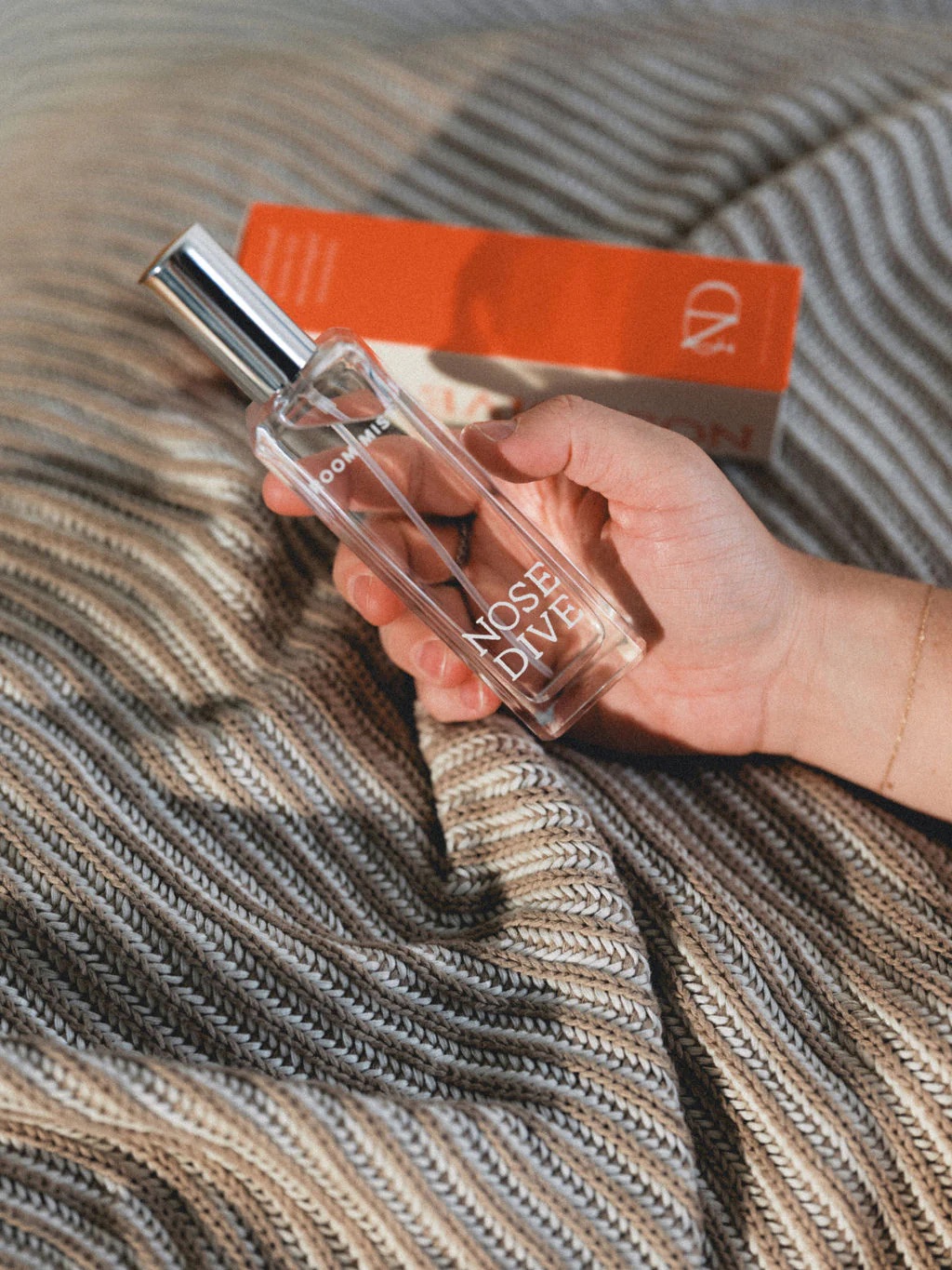 A hand holds a clear perfume bottle labeled "NOSE DIVE" above a striped knit blanket, with an orange and white box in the background.