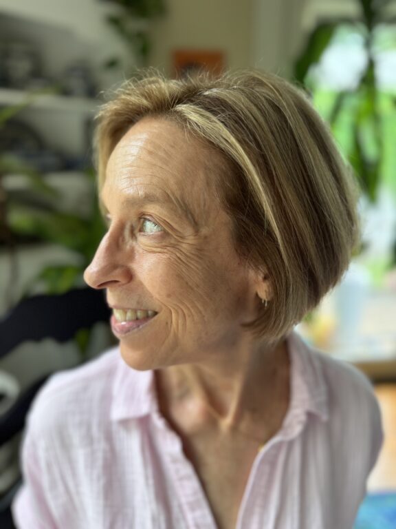 A woman with short blonde hair and a light pink shirt looks to her left, smiling. The background shows indoor plants and shelves.