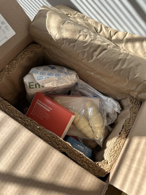 A cardboard box with paper padding contains packaged food items, including a vacuum-sealed bag, a red booklet, and a bag of peeled potatoes.