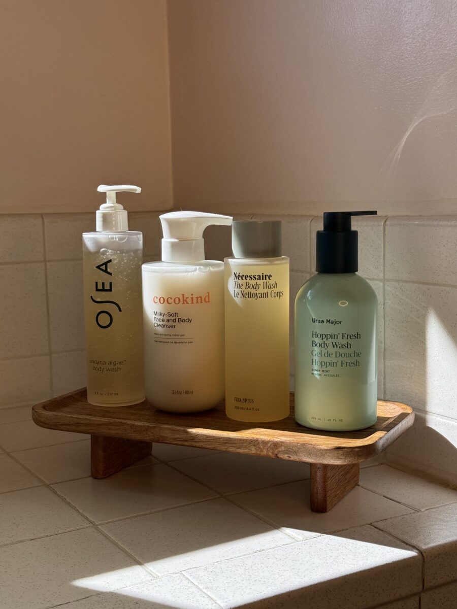 Four bottles of body wash and lotion are arranged on a wooden shelf in a tiled bathroom corner, lit by natural sunlight.