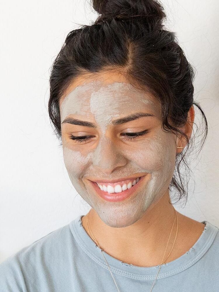 A woman with dark hair smiles while wearing a gray facial mask and a light blue shirt, standing against a plain white background.