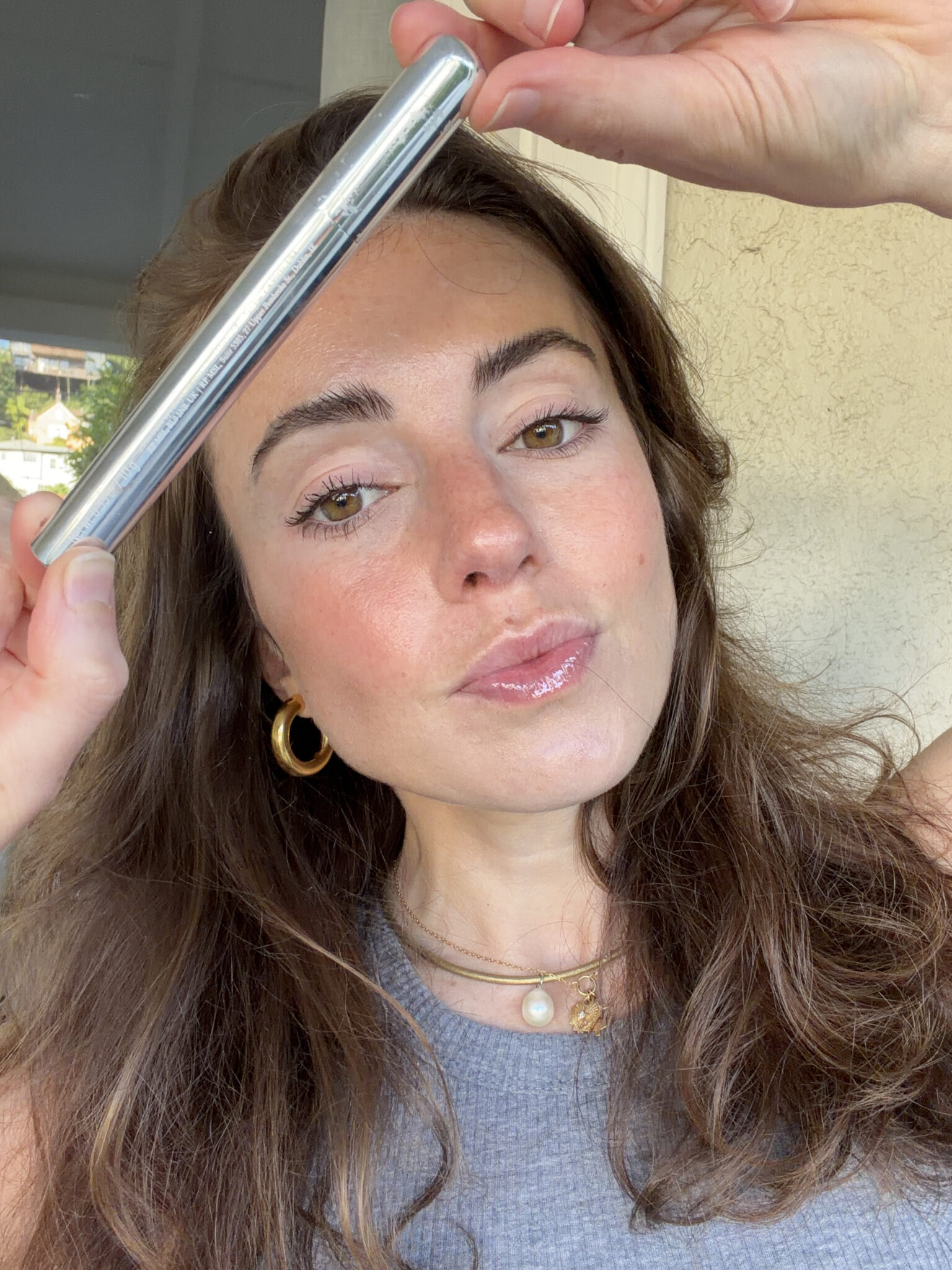 A woman with long brown hair holds a silver cylindrical object above her forehead while looking at the camera. She wears gold hoop earrings and a gray top.