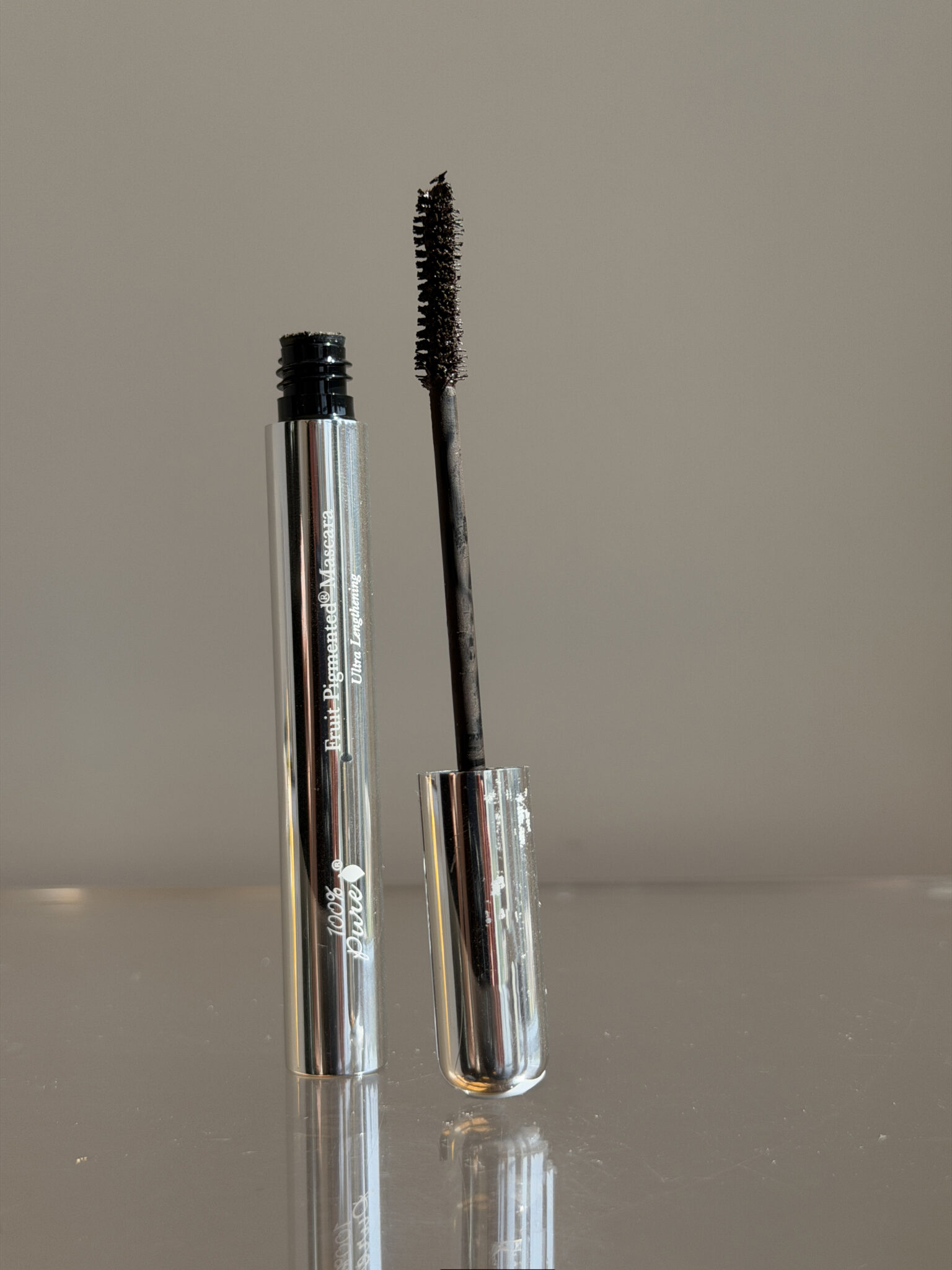 A silver tube of mascara stands upright with its wand and brush exposed, coated in black mascara, against a neutral background.