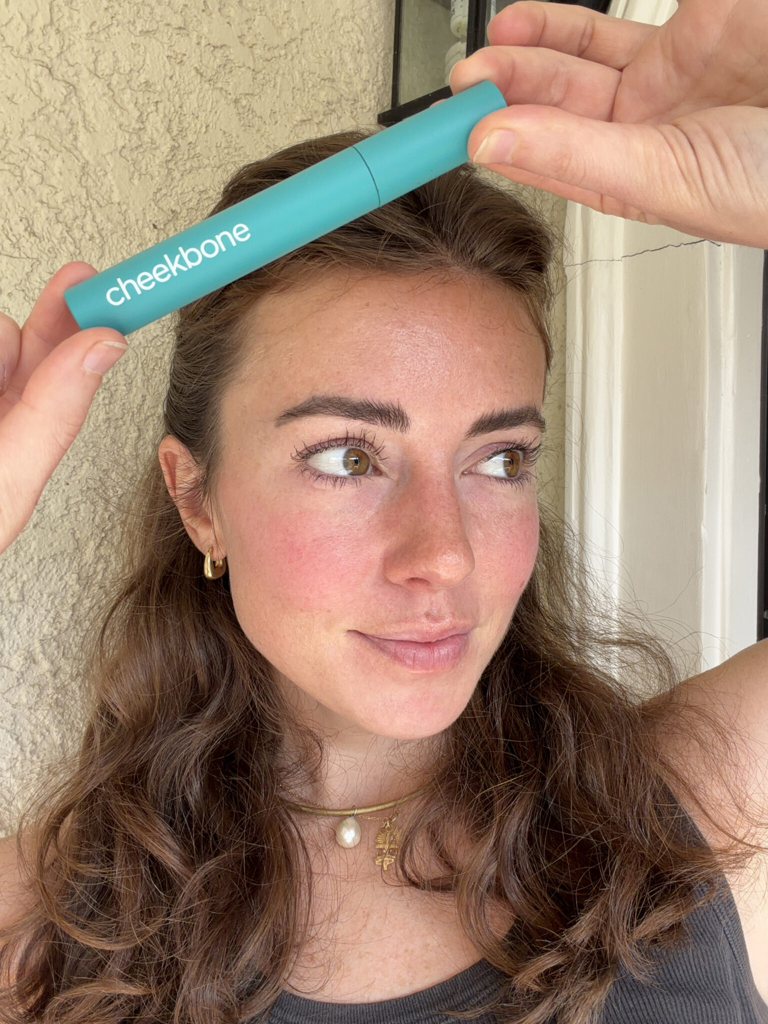 A woman with long brown hair holds a turquoise Cheekbone mascara tube above her forehead while standing in front of a beige wall.