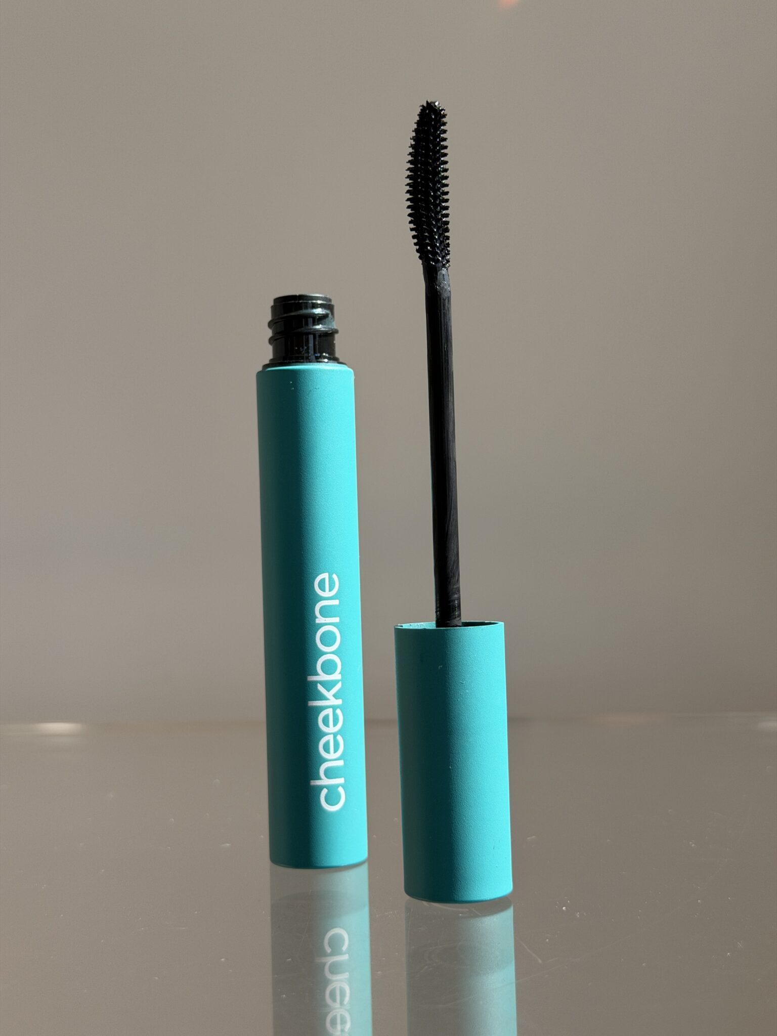 A teal tube of cheekbone mascara stands upright with the wand applicator removed and resting vertically beside it on a reflective surface.
