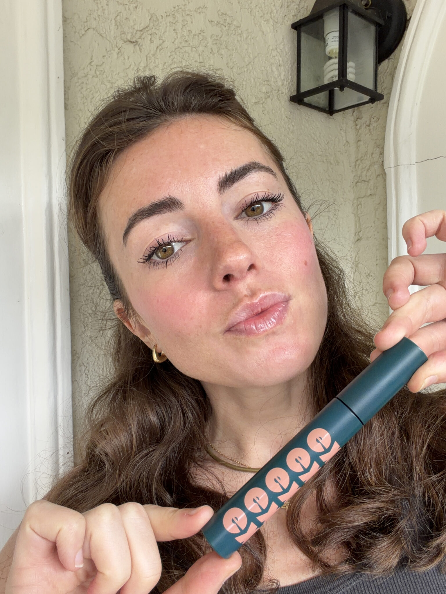A woman with long brown hair holds a green tube of mascara, showing her eyelashes and looking at the camera.