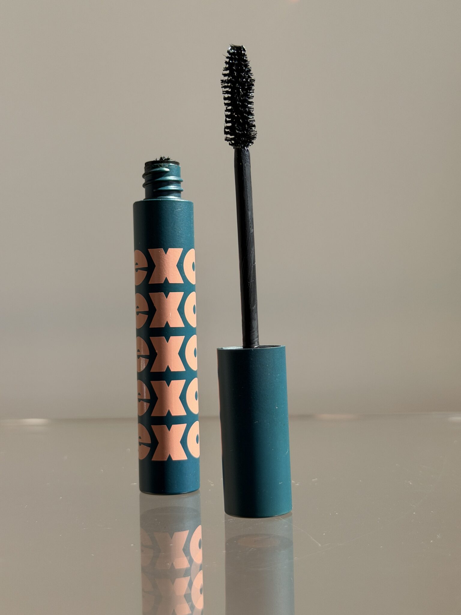 An open tube of mascara with a teal and orange design, showing the brush applicator standing upright next to the container on a reflective surface.
