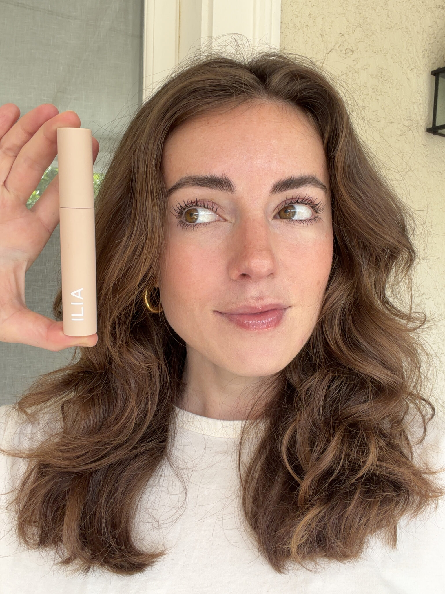 A woman with wavy brown hair holds a beige ILIA makeup product next to her face while looking at it. She is wearing a white top and standing in front of a neutral background.