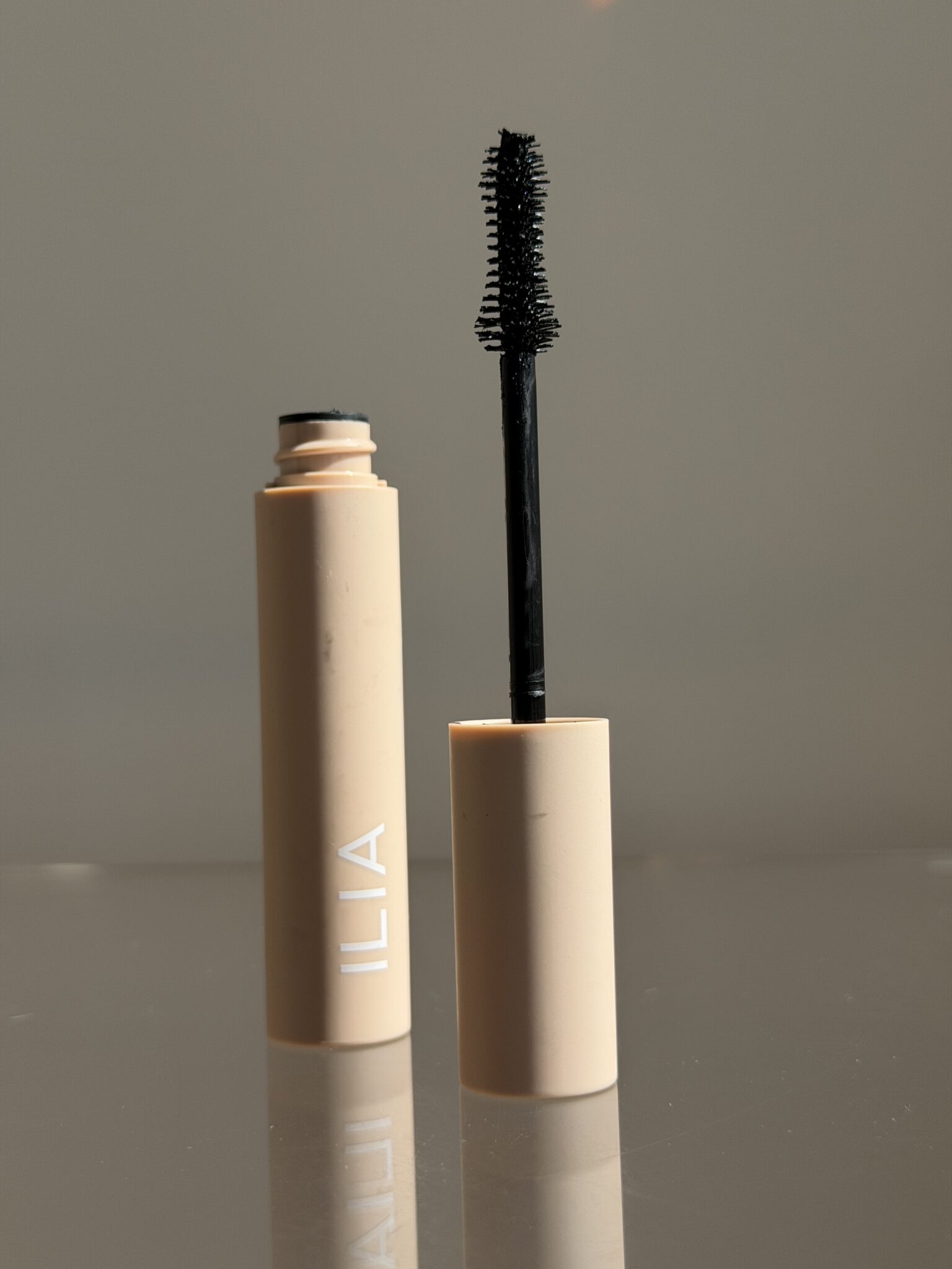 A tube of ILIA mascara with the wand applicator removed and standing upright in the tube, displayed on a reflective surface.