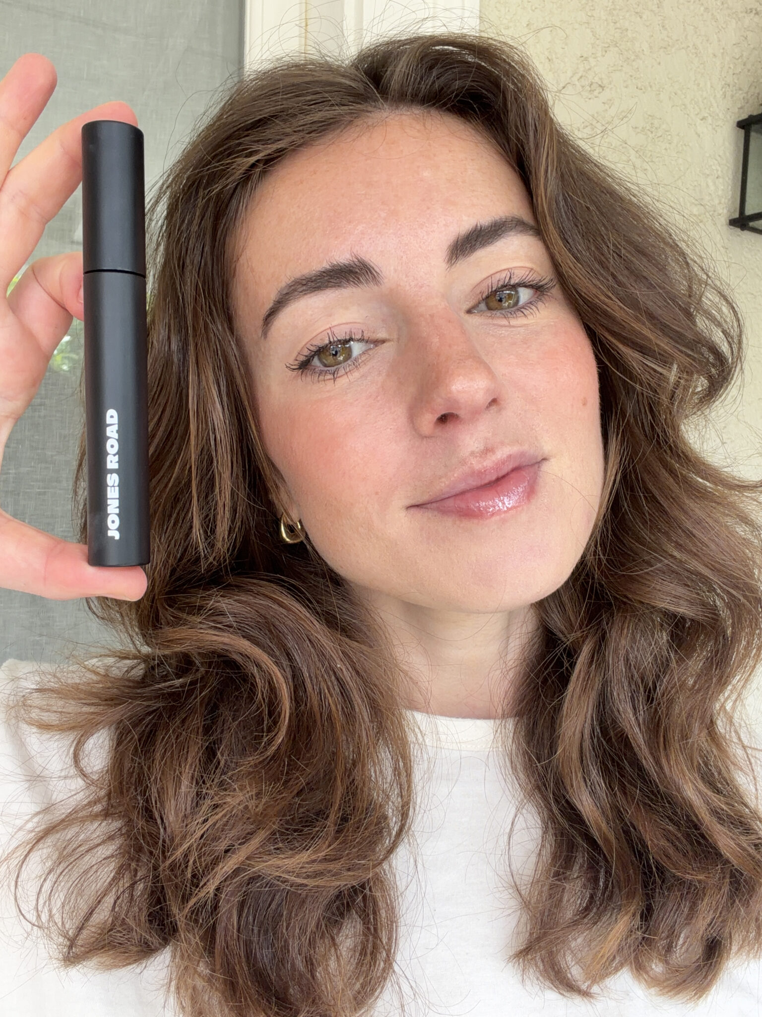 A woman with wavy brown hair holds up a black tube of Jones Road mascara while looking at the camera.