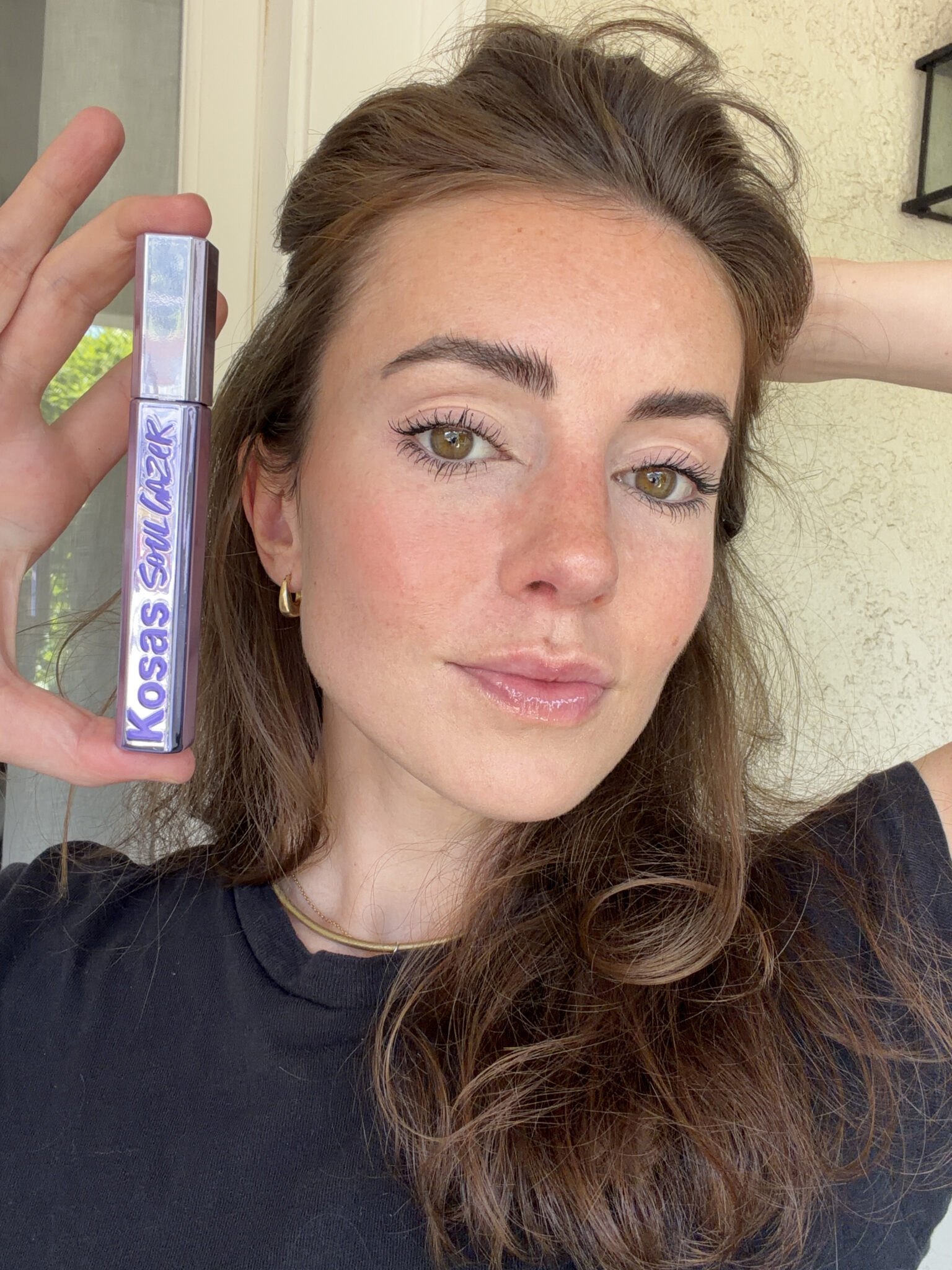 A woman with long brown hair holds up a tube of Kosas mascara next to her face, showing her natural makeup look.
