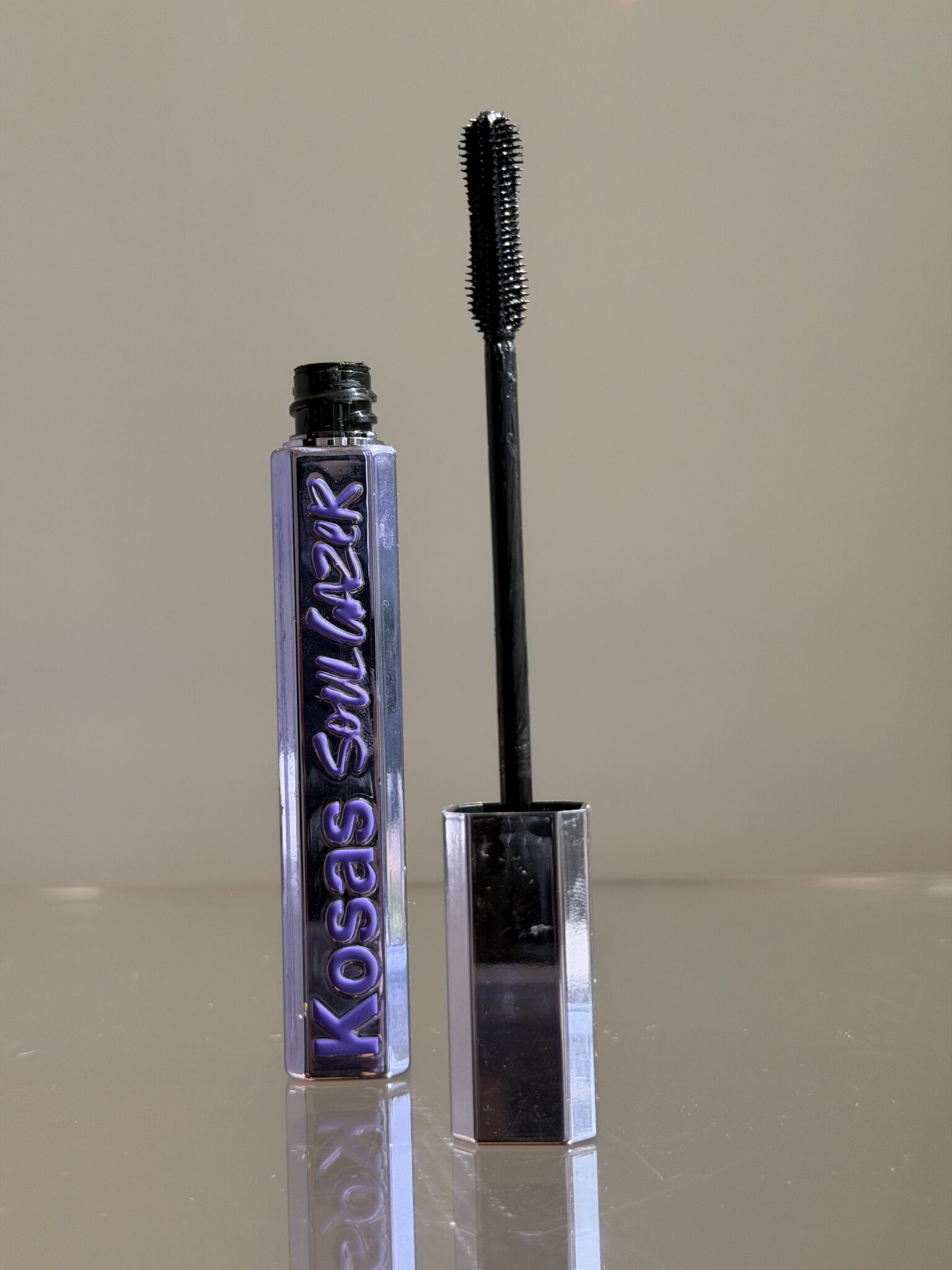 A tube of Kosas mascara with the wand standing upright beside it, both on a reflective surface against a neutral background.