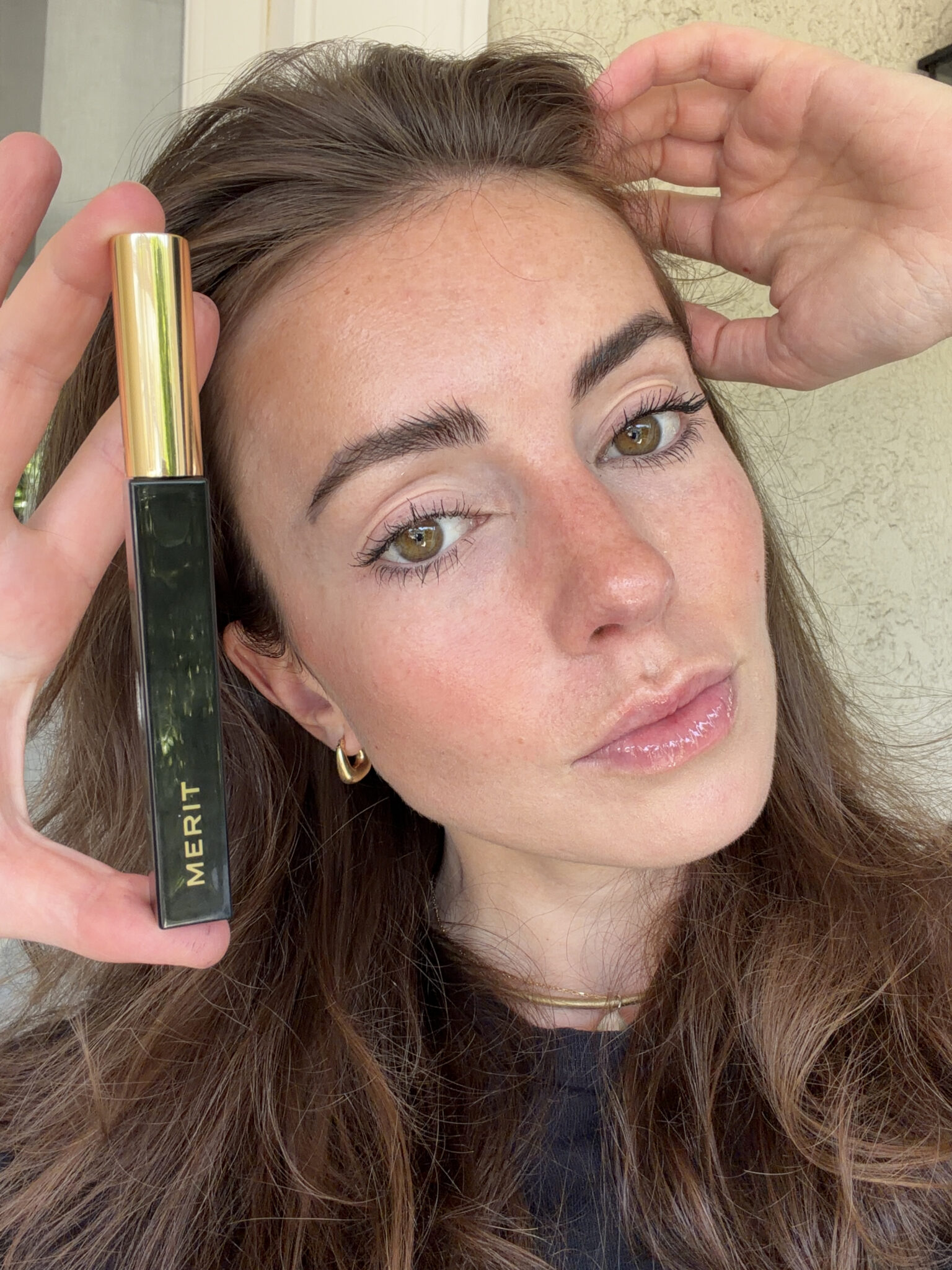 A woman with long brown hair holds up a black tube of Merit mascara next to her face, showcasing her natural makeup look.