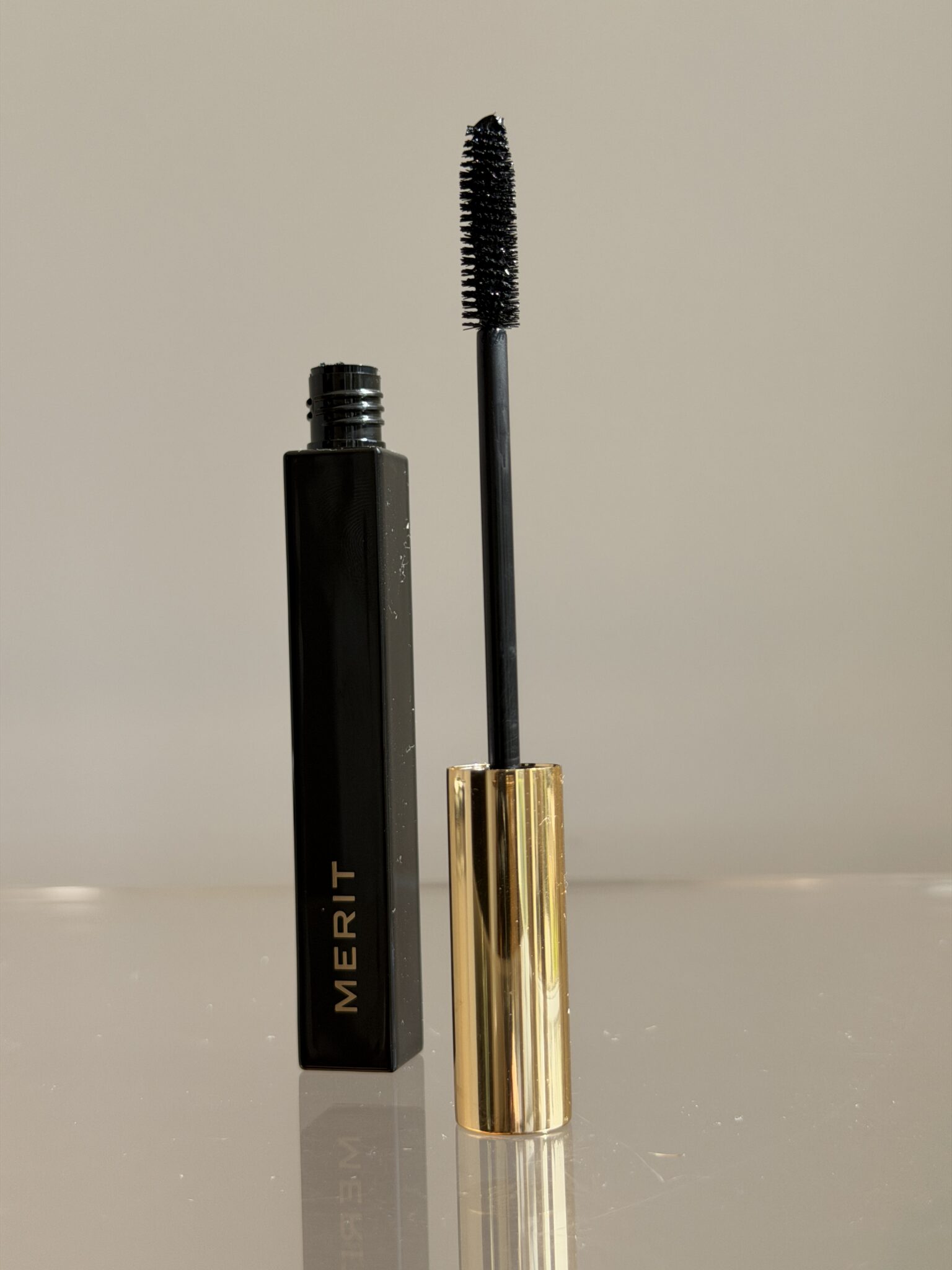 A black tube of MERIT mascara stands upright with its gold cap removed and attached to the wand, which is coated with black mascara.