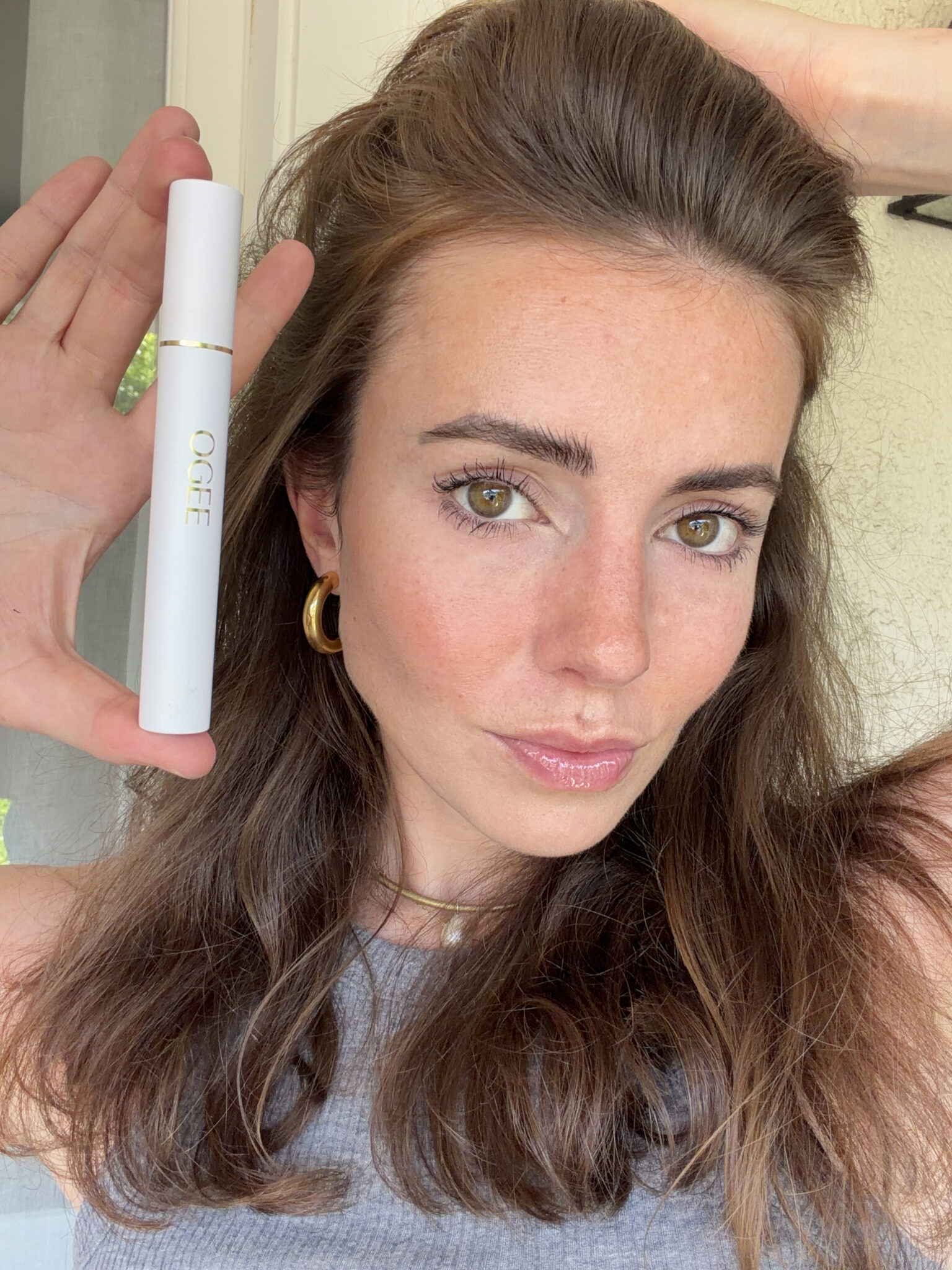 A woman with long brown hair holds up a white tube labeled "OGEE" next to her face, posing for a close-up photo indoors.