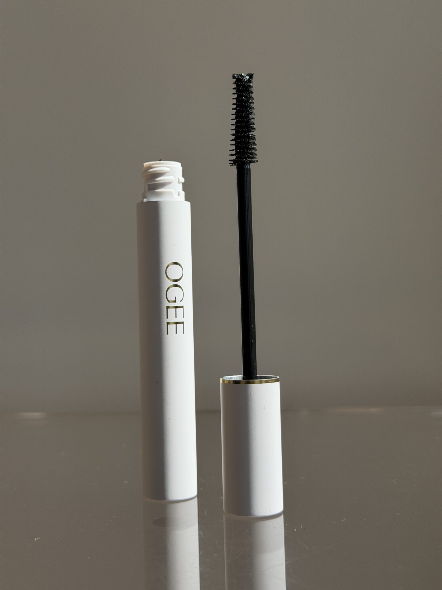 A white tube of OGEE mascara with the wand removed and standing upright beside the open container against a neutral background.