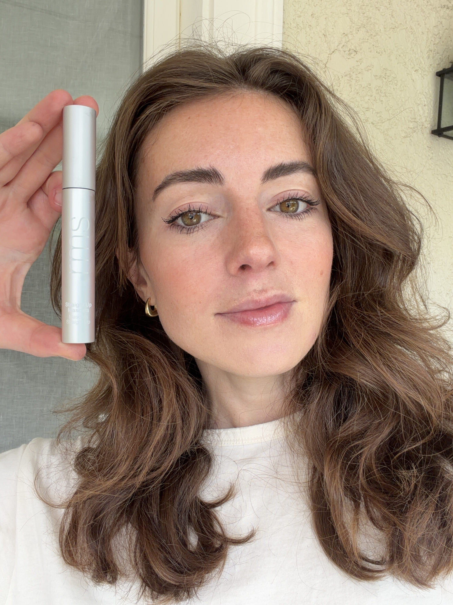 A woman with wavy brown hair holds up a silver tube of Surratt mascara next to her face, standing indoors near a beige wall.