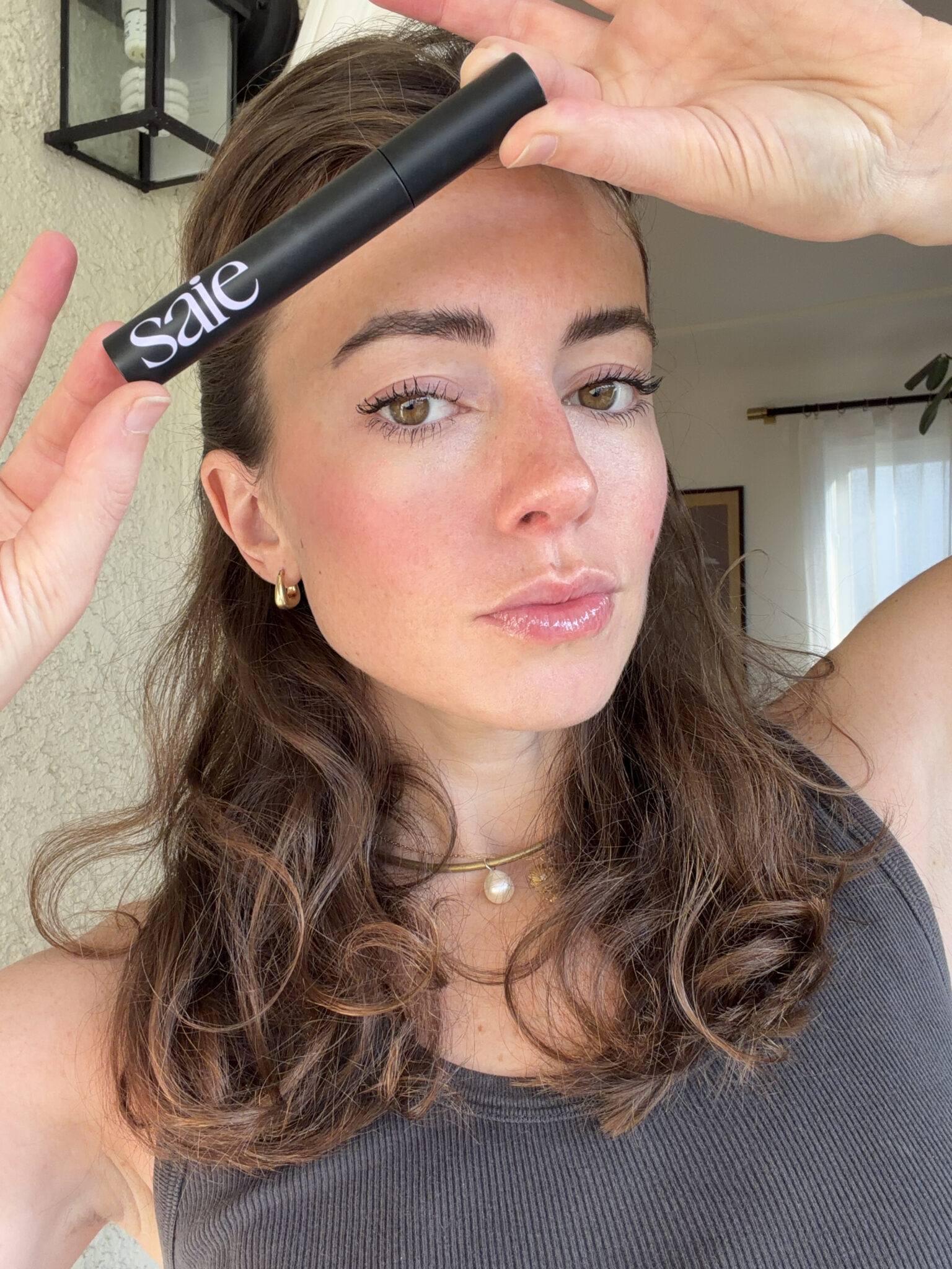 A woman holds a black tube of mascara labeled "saie" above her forehead, wearing a dark tank top and gold jewelry, with natural makeup and wavy brown hair.