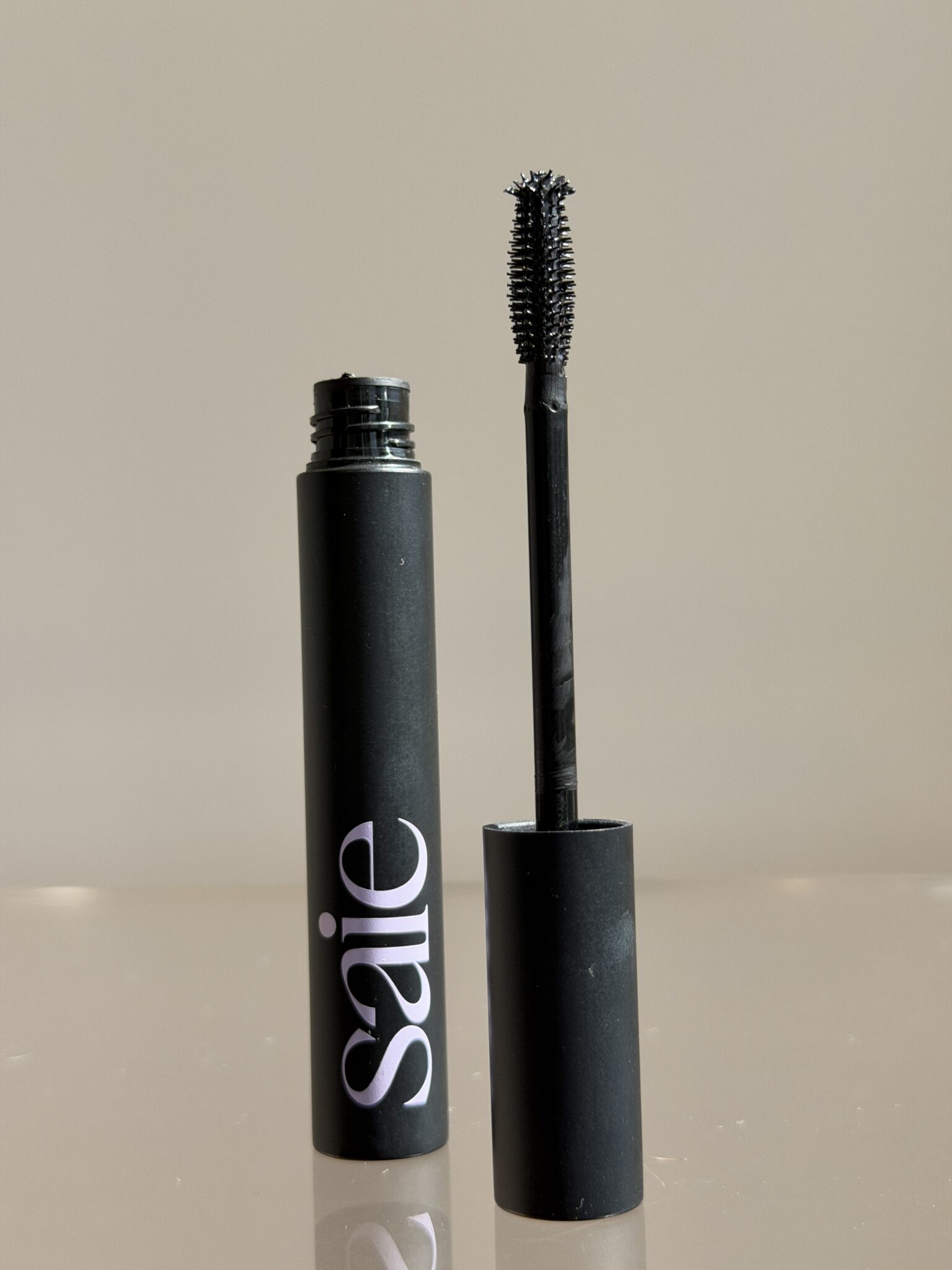 A black tube of Saie mascara with the wand removed and standing upright beside the open container against a neutral background.