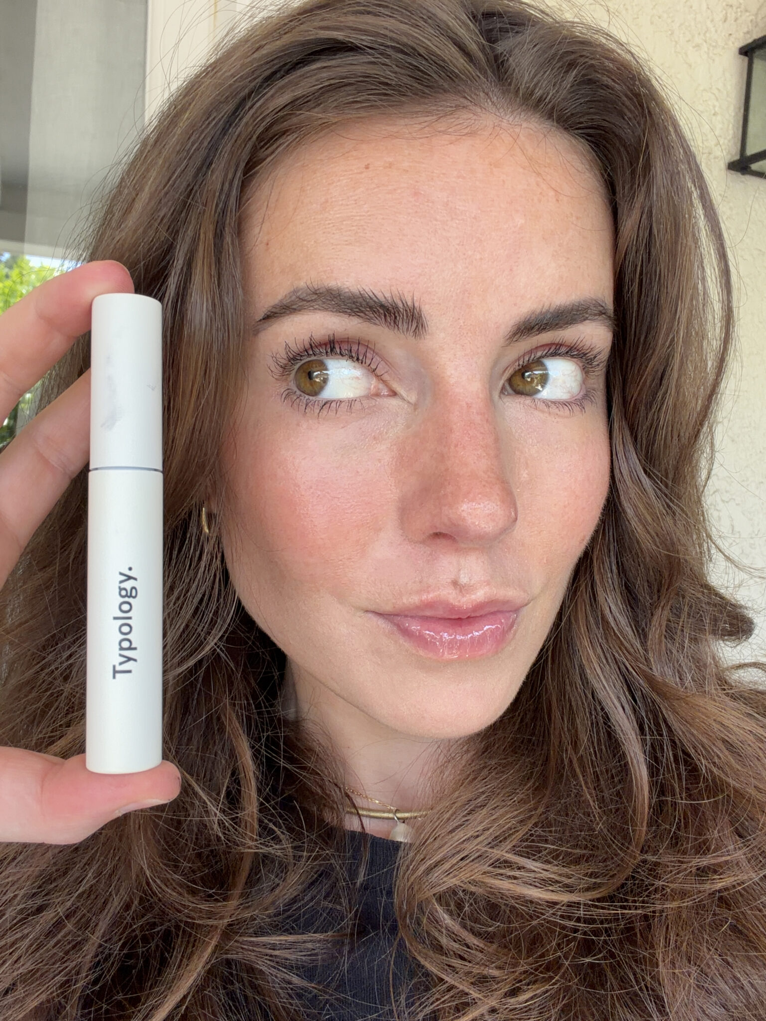 Woman with wavy brown hair holds a Typology product next to her face, looking slightly to the side with a neutral expression.