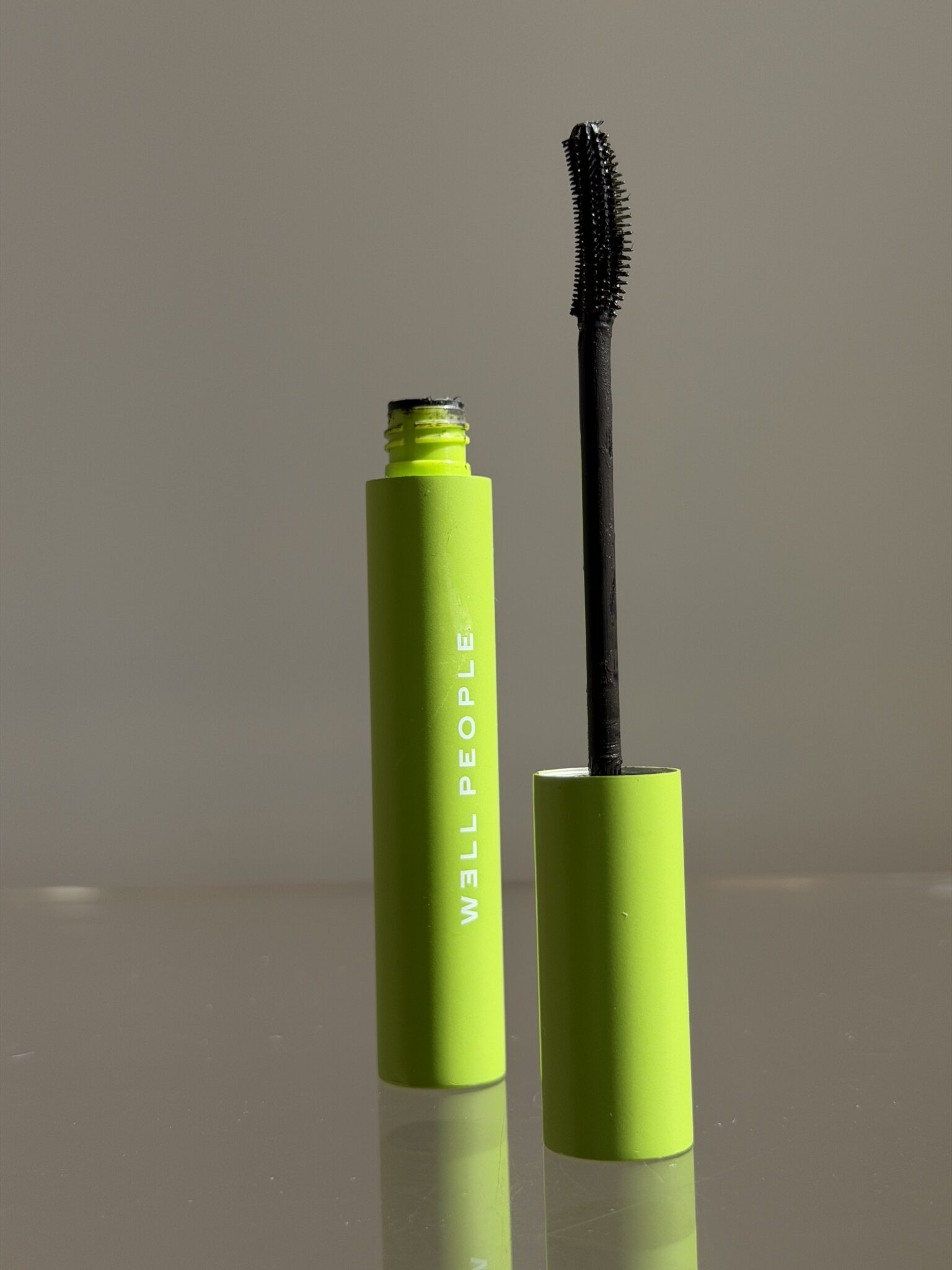 A green tube of WELL PEOPLE mascara with the wand applicator removed and standing upright beside it against a neutral background.