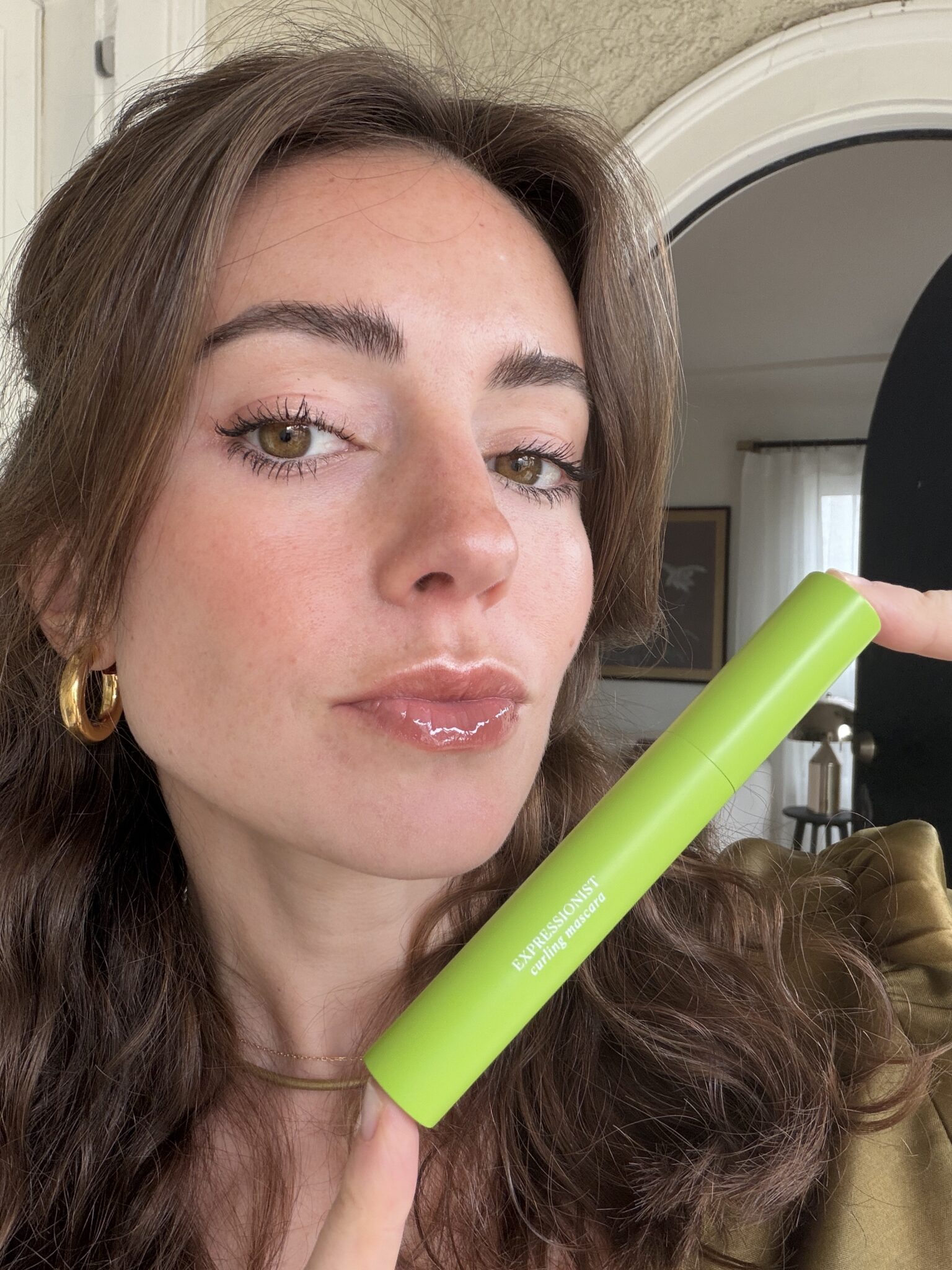 A woman with wavy brown hair holds up a green tube of mascara next to her face, showing her glossy lips and defined eyelashes.
