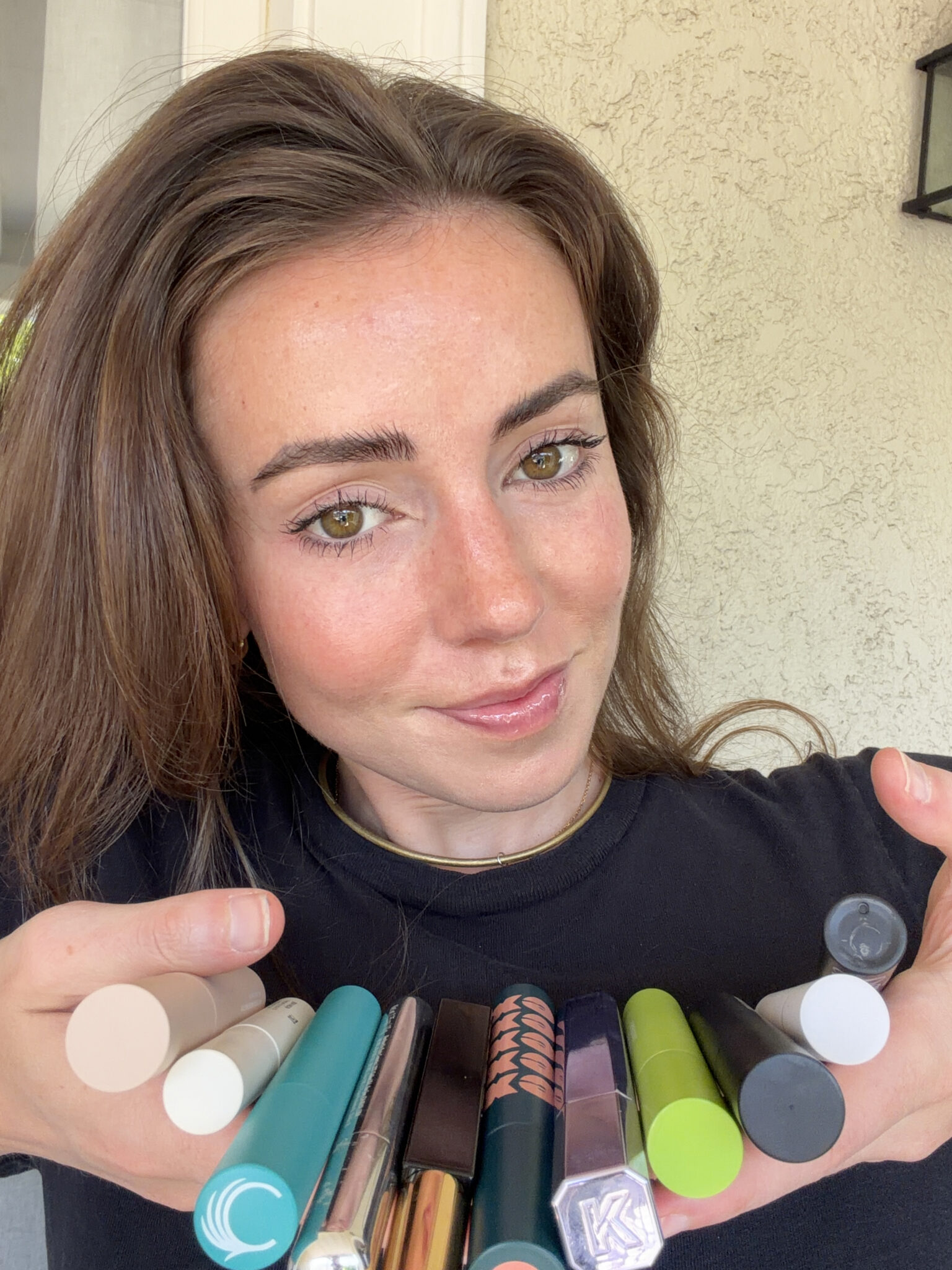 A woman holds an array of colorful mascara tubes in both hands, displaying them in front of her chest while smiling at the camera.