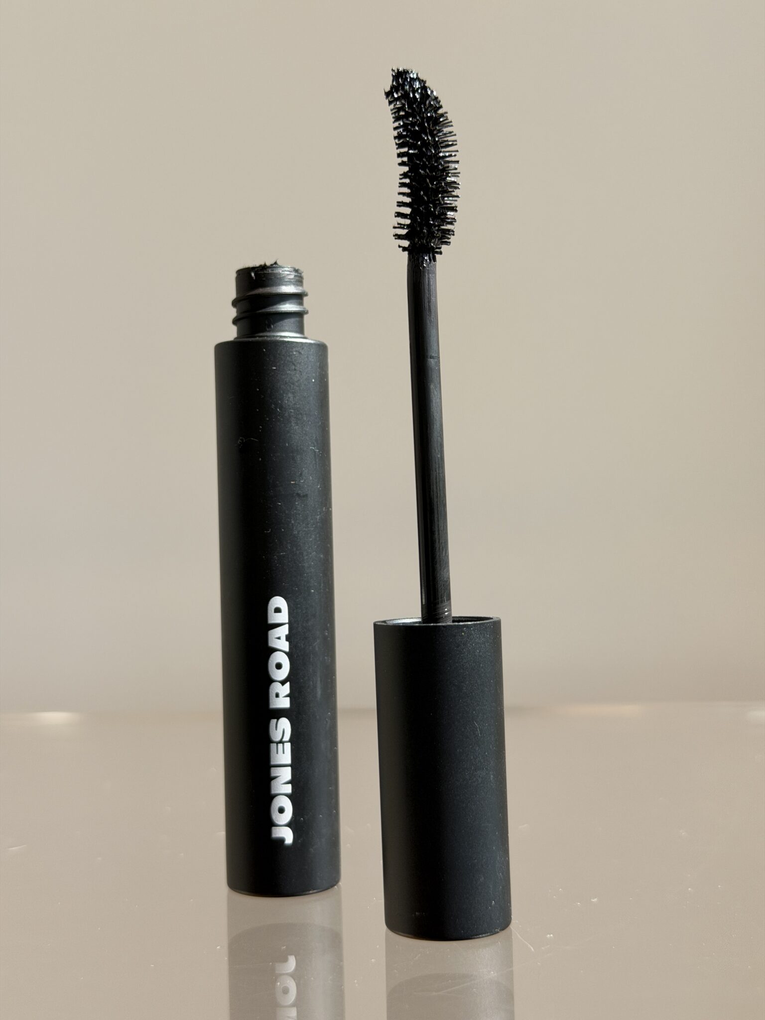 A black tube of Jones Road mascara stands upright with the wand removed, showing a curved brush coated in black mascara against a neutral background.