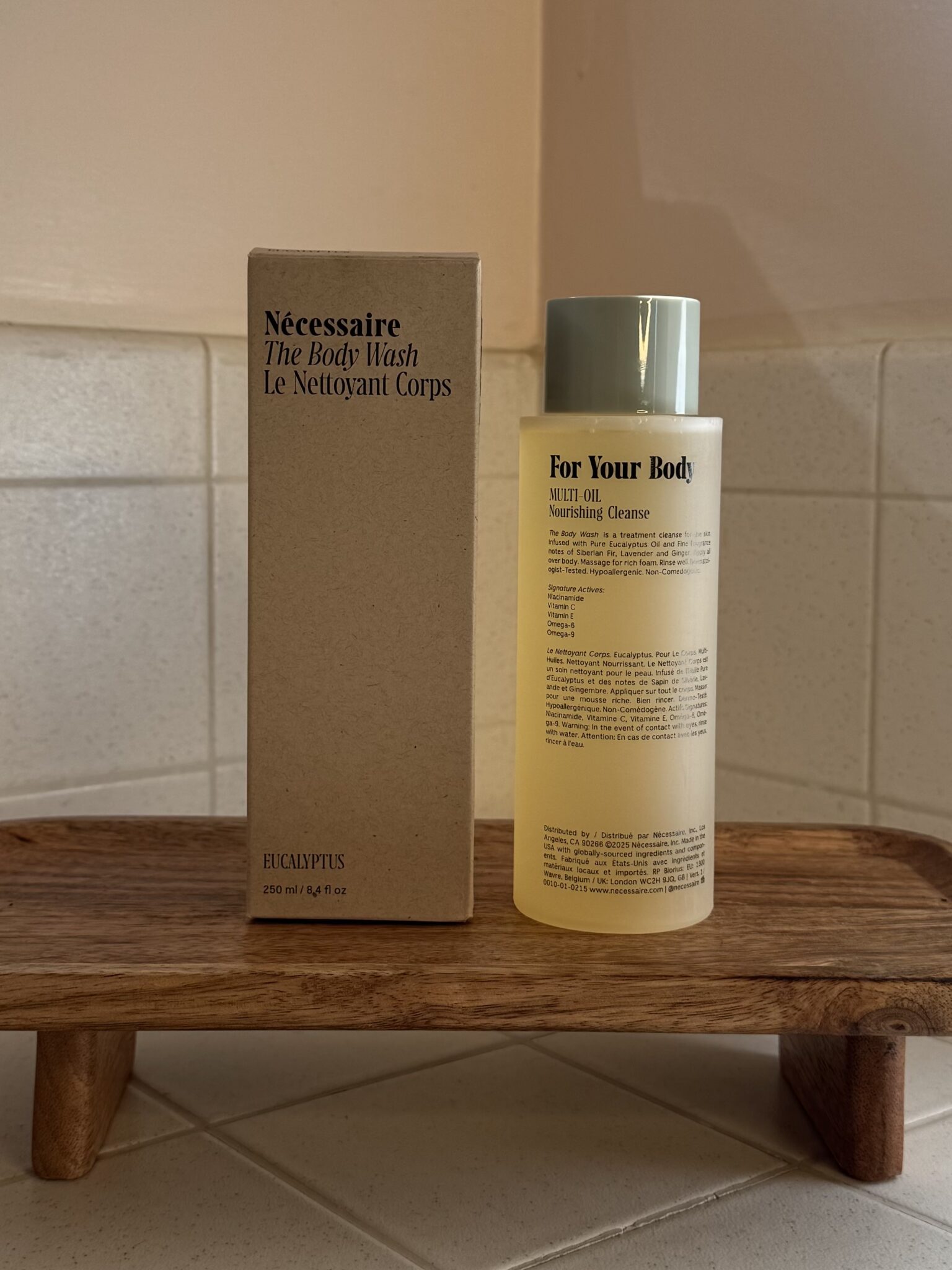 A bottle of body wash labeled "For Your Body" and a box labeled "Nécessaire The Body Wash" are placed on a wooden tray in a tiled bathroom setting.
