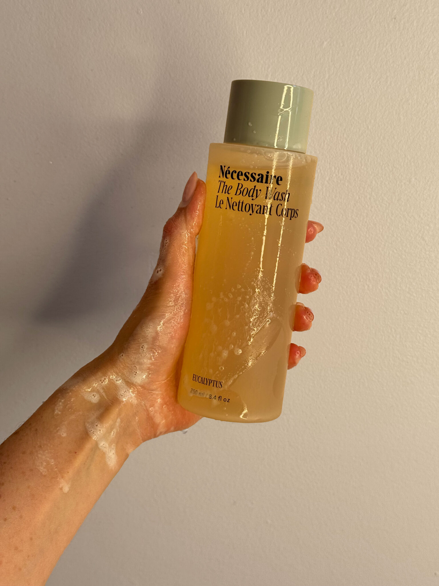 A hand holding a bottle of Nécessaire The Body Wash in eucalyptus scent, with soap suds visible on the hand and bottle against a plain background.