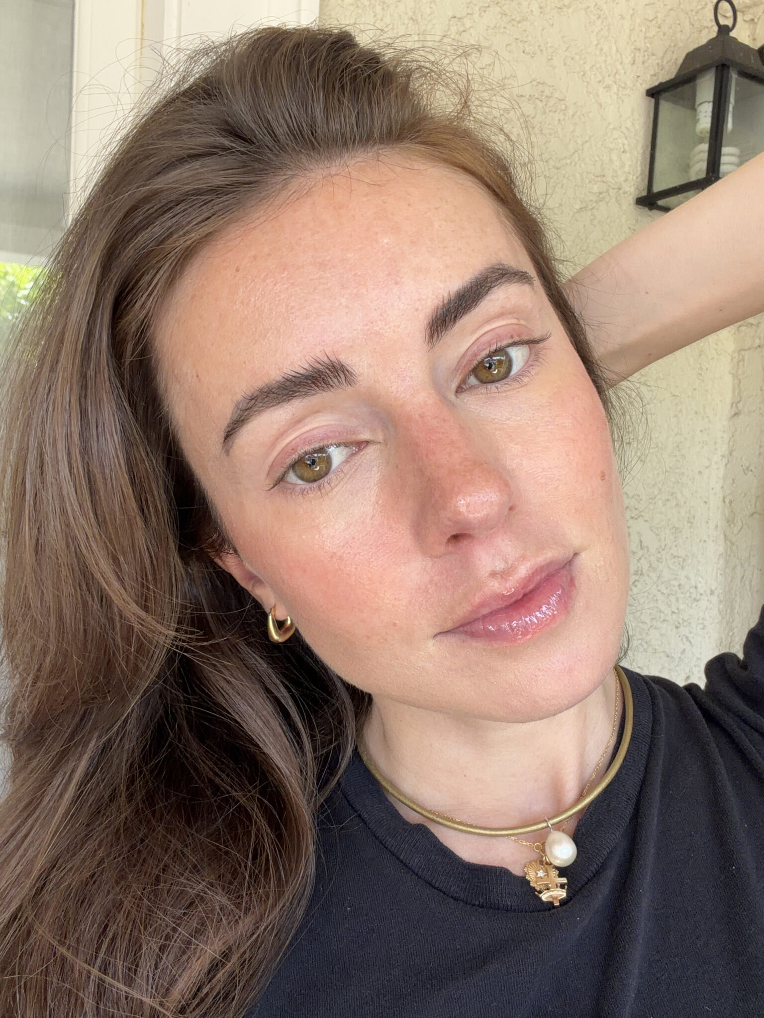 A woman with long brown hair poses indoors, wearing a black top, gold hoop earrings, and a gold necklace with a pearl pendant. She has a neutral facial expression.
