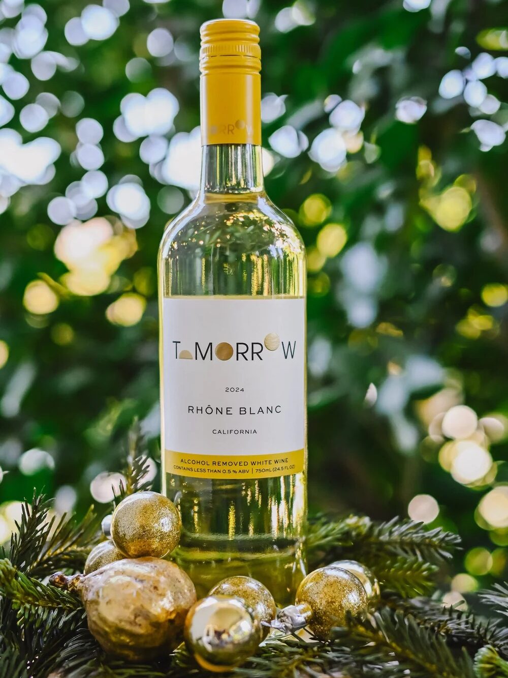 A bottle of Tomorrow 2022 Rhône Blanc white wine stands on pine branches with gold ornaments, set against a blurred green foliage background.