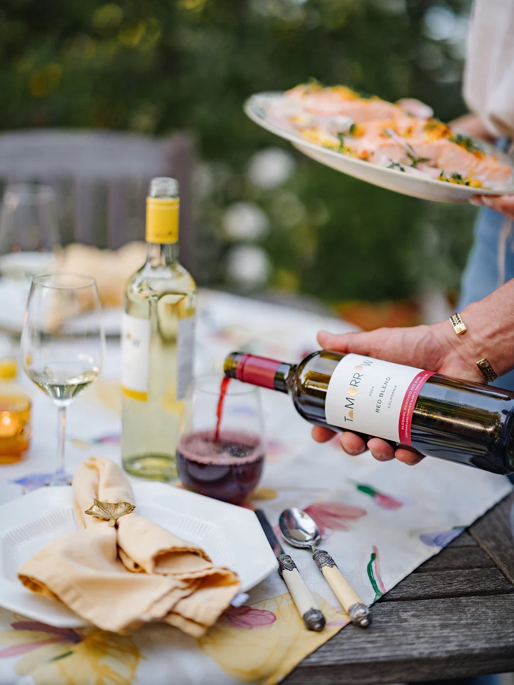 A person pours red wine into a glass on a table set with two wine bottles, an empty glass, plates, silverware, and a platter of food.