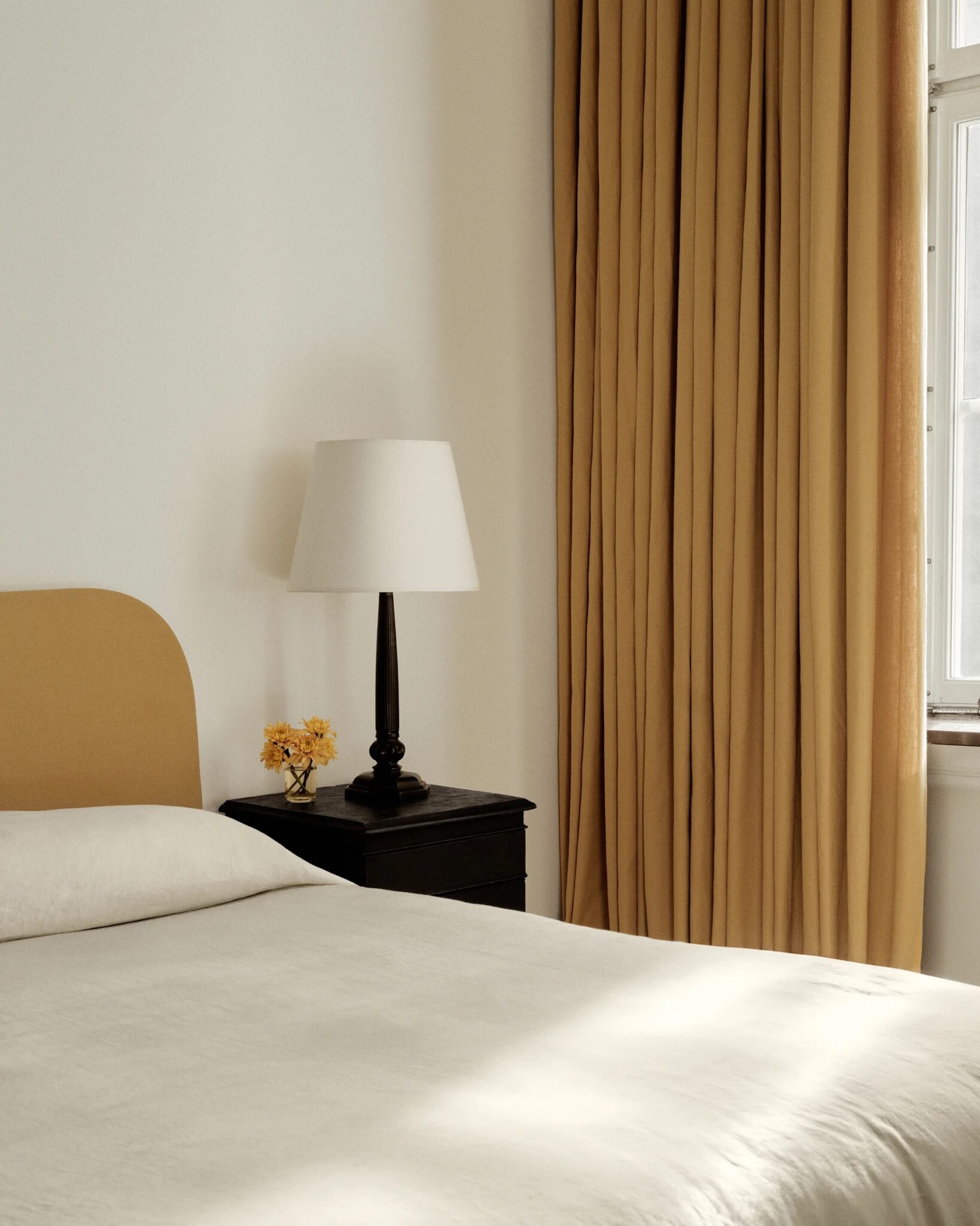 A neatly made bed with a beige headboard, a black nightstand with a lamp and small yellow flowers, and beige curtains beside a window letting in natural light.