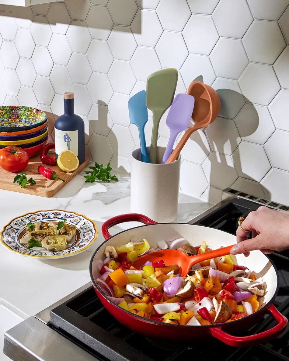 A person stirs chopped vegetables in a red skillet on a stove; utensils are in a holder nearby and fresh ingredients sit on a cutting board.