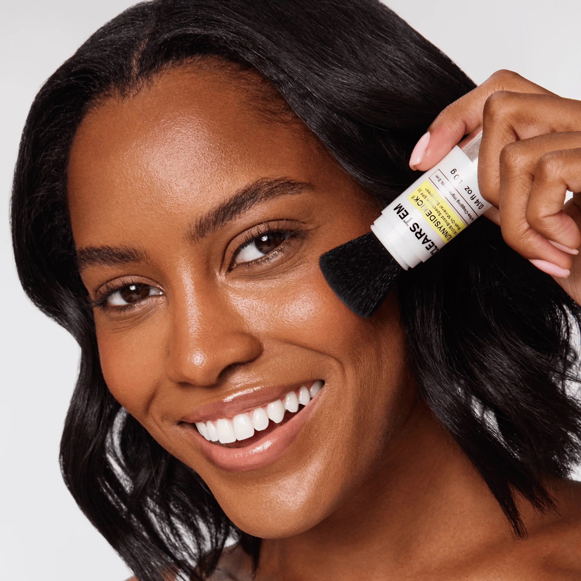 A person smiles while applying a stick product to their cheek with a built-in brush.