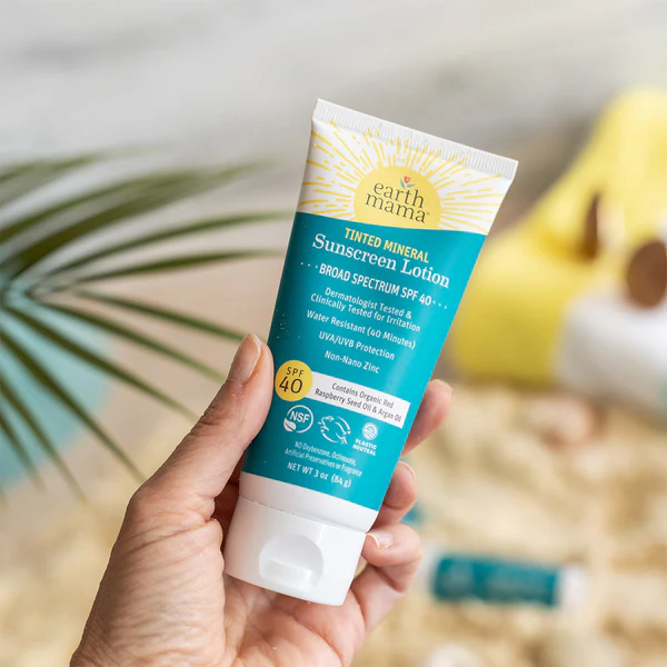 A hand holds a tube of Earth Mama Tinted Mineral Sunscreen Lotion SPF 40; blurred beach items are in the background.