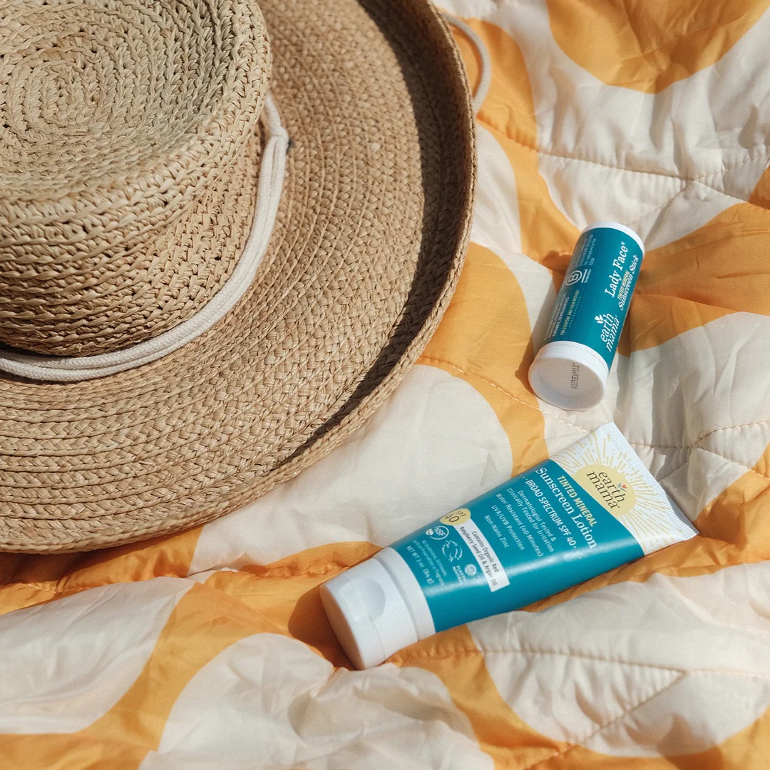 A straw hat, a tube of sunscreen, and a stick sunscreen are placed on a yellow and white patterned blanket.