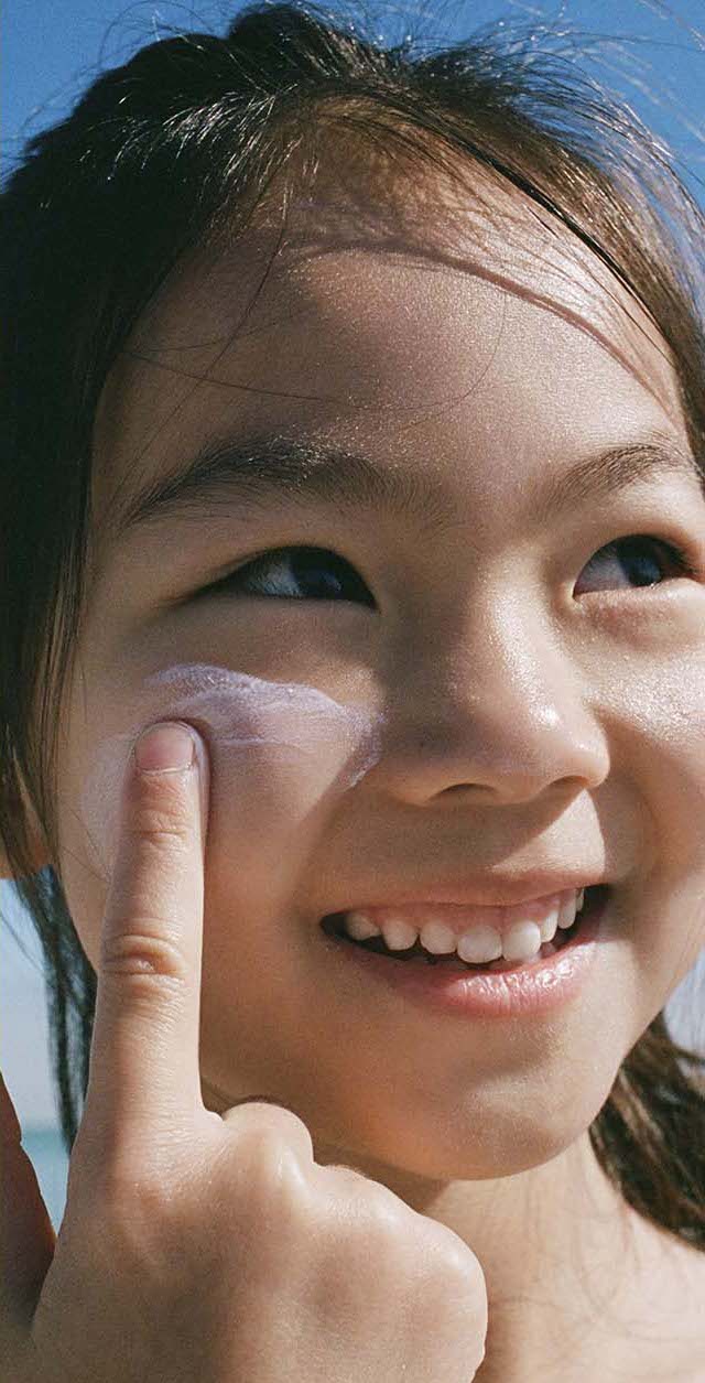 A smiling child applies sunscreen to their face with a finger under a bright blue sky.