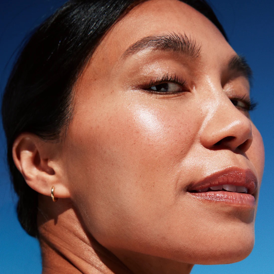 Close-up of a woman’s face with clear skin, minimal makeup, and a small gold hoop earring, set against a bright blue sky.