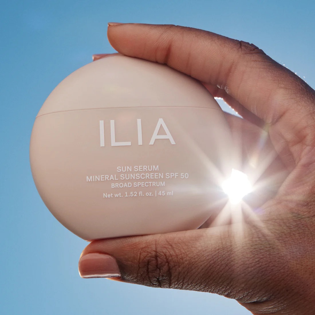 A hand holds a round container of ILIA Sun Serum Mineral Sunscreen SPF 50 against a blue sky with sunlight shining through the fingers.