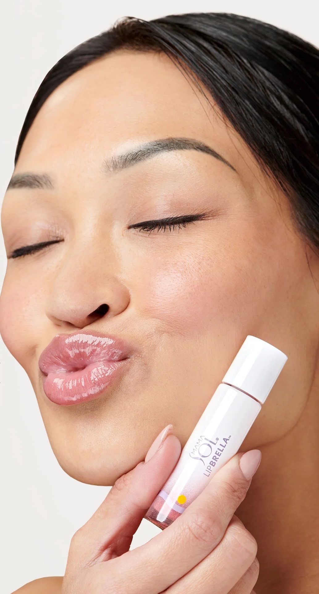 A person with closed eyes puckers their lips and holds a tube of clear lip gloss labeled "SOL by Jergens" near their face.