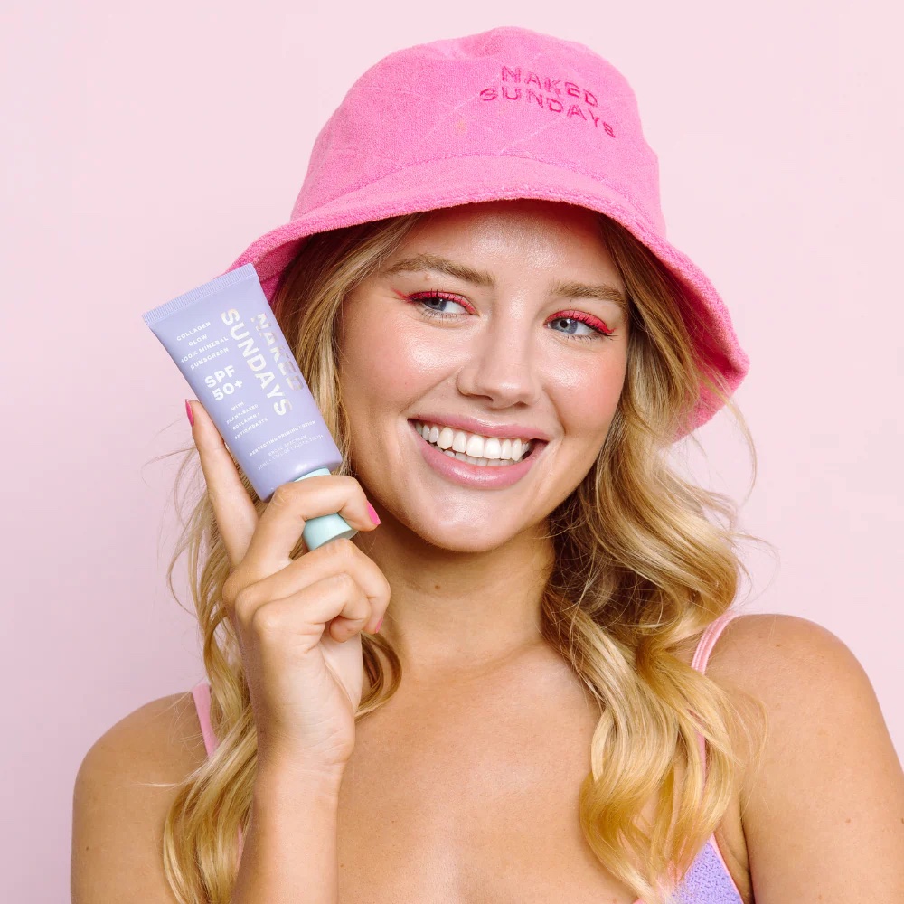 A woman in a pink hat holds up a tube of Sunscreen SPF 50+ against a pink background, smiling.
