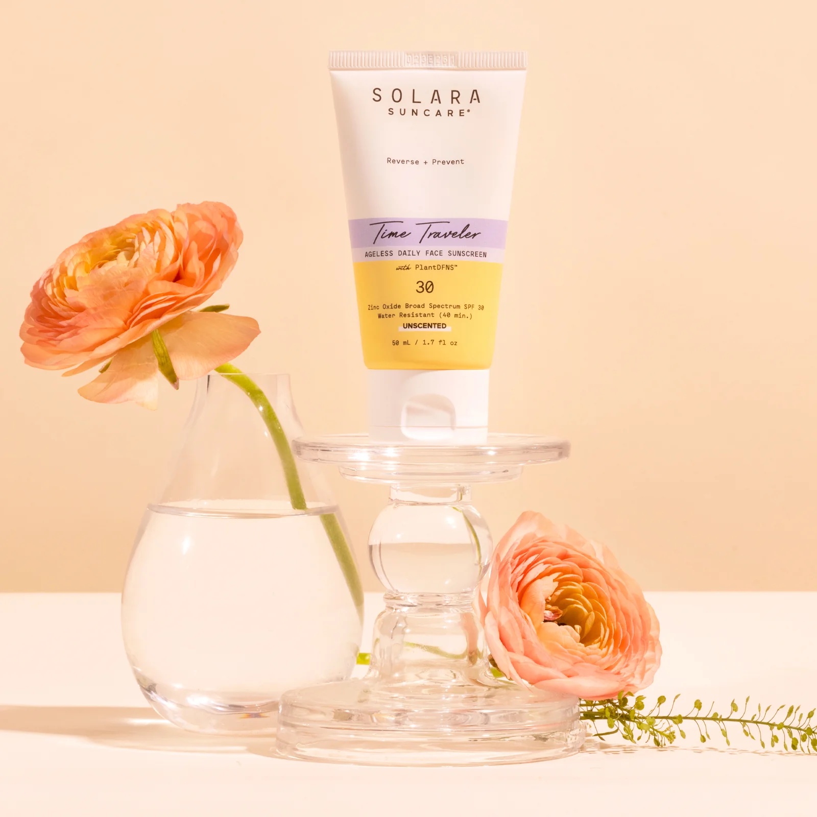 A tube of Solara Suncare sunscreen is displayed on a glass stand next to two orange ranunculus flowers in a glass of water against a beige background.