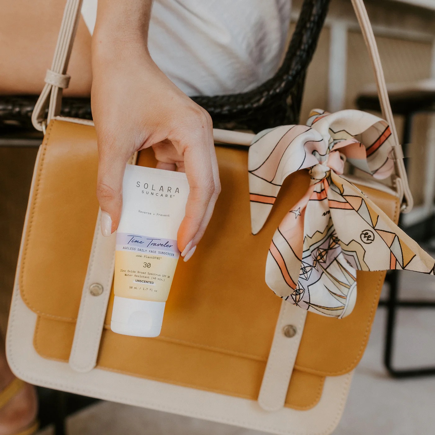 A person holds a tube of Solara Suncare sunscreen next to a tan handbag with a patterned scarf tied to the handle.