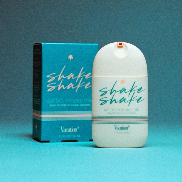 A bottle and box of Vacation brand Shake Shake SPF 50 mineral milk sunscreen are displayed against a blue background.
