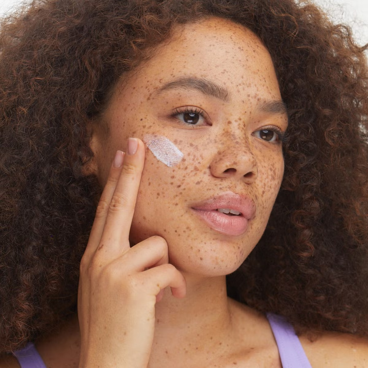 A woman with curly hair and freckles applies a dab of cream to her cheek with her fingertip.