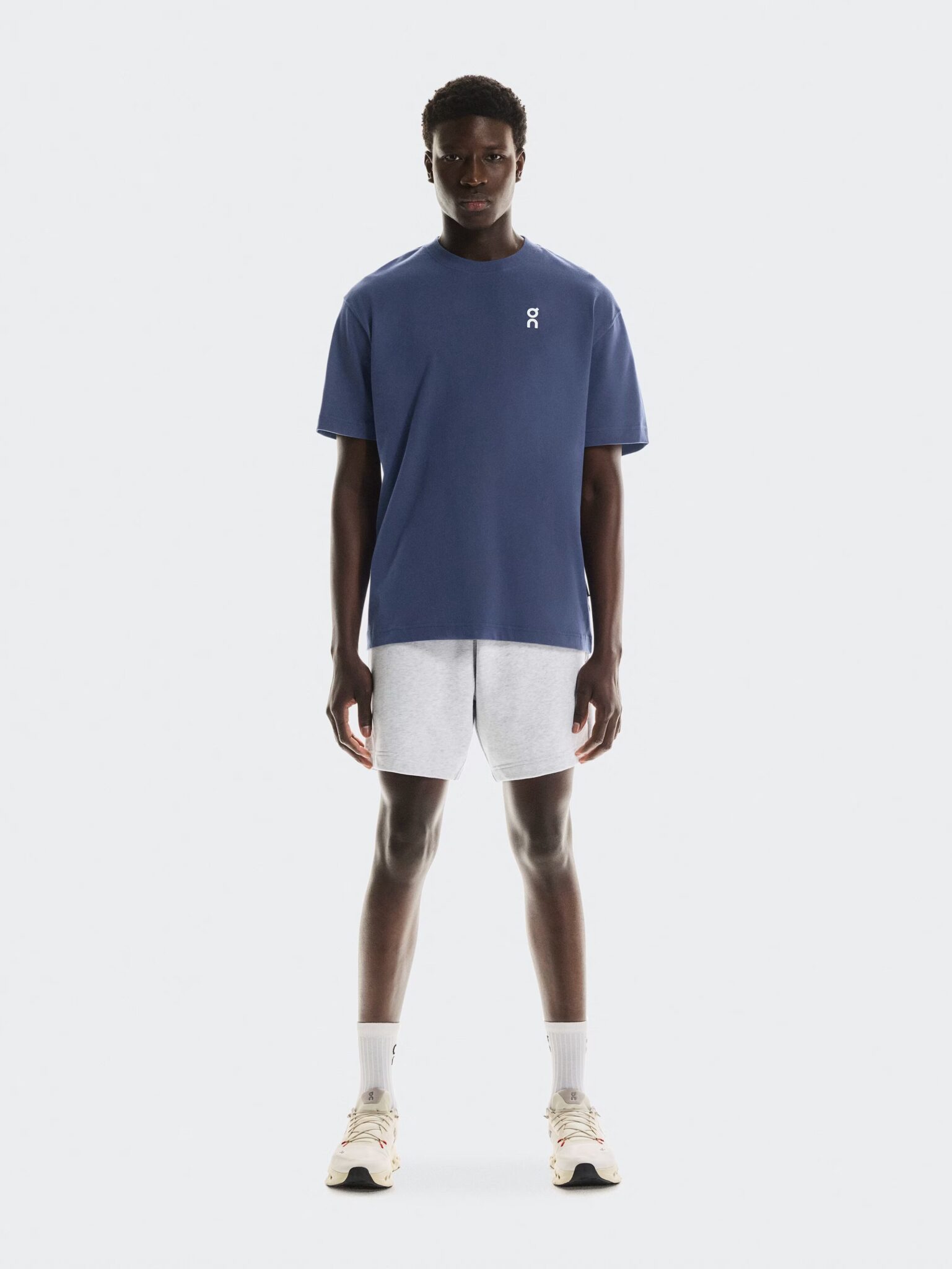 A person stands facing forward wearing a blue t-shirt, light gray shorts, white socks, and white sneakers against a plain white background.